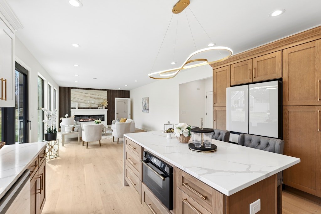 Modern open-concept kitchen and living room with white walls, light wood flooring, natural wood cabinets, white marble countertop, black and white refrigerator, gray upholstered chairs, white seating area with a fireplace, and large windows with slid