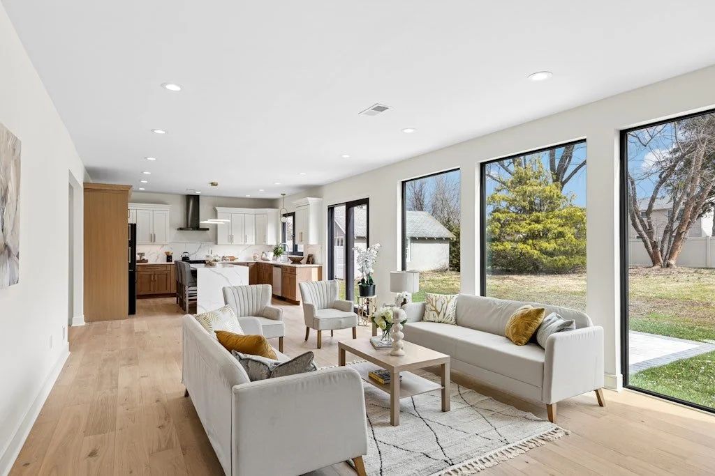 Bright living room with white sofas, wooden coffee table, large sliding glass doors, and views of a backyard with grass and trees.