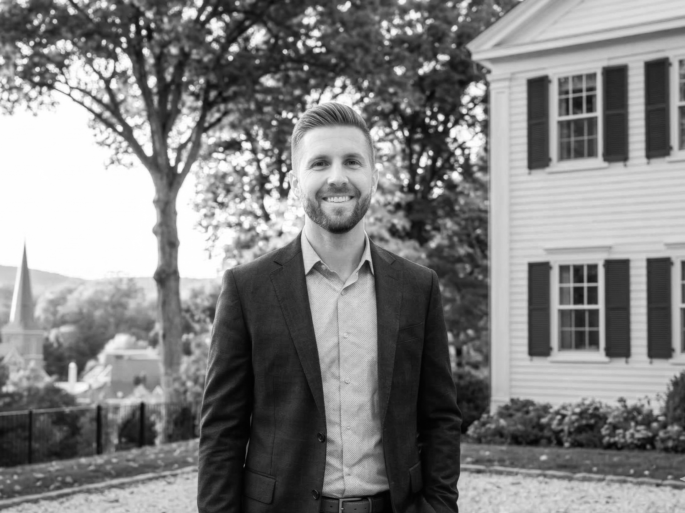 A smiling man in a suit standing outdoors near a white house with green shutters, trees with fall foliage in the background.