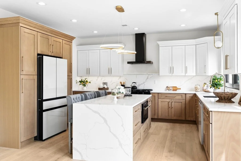 Modern kitchen with white and light wood cabinets, marble countertops, and a large island with seating. Appliances include a black range, a black and white fridge, and a microwave. Decor features plants, a woven basket, and flowers, with a contempora