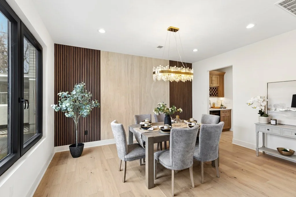Modern dining room with a wooden table, six gray upholstered chairs, a chandelier, and decorative plants, with a window and a small kitchen area visible.
