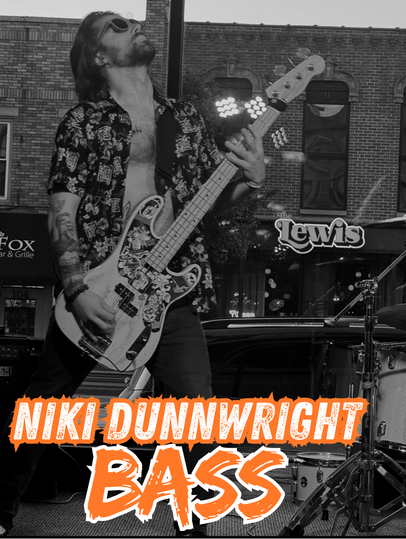 A man with long hair, wearing sunglasses and a floral shirt, playing an electric bass guitar on stage, with a drummer and a brick building in the background.