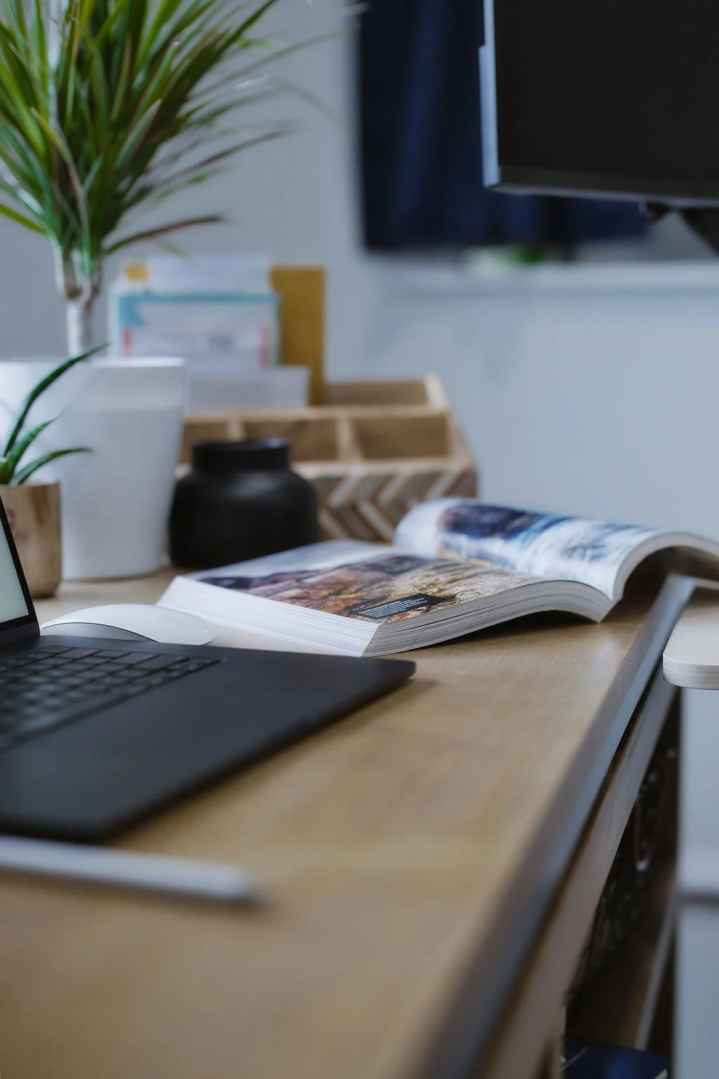A desk with an open magazine, a laptop, potted plants, and office supplies, with a blurred background of a window and storage.