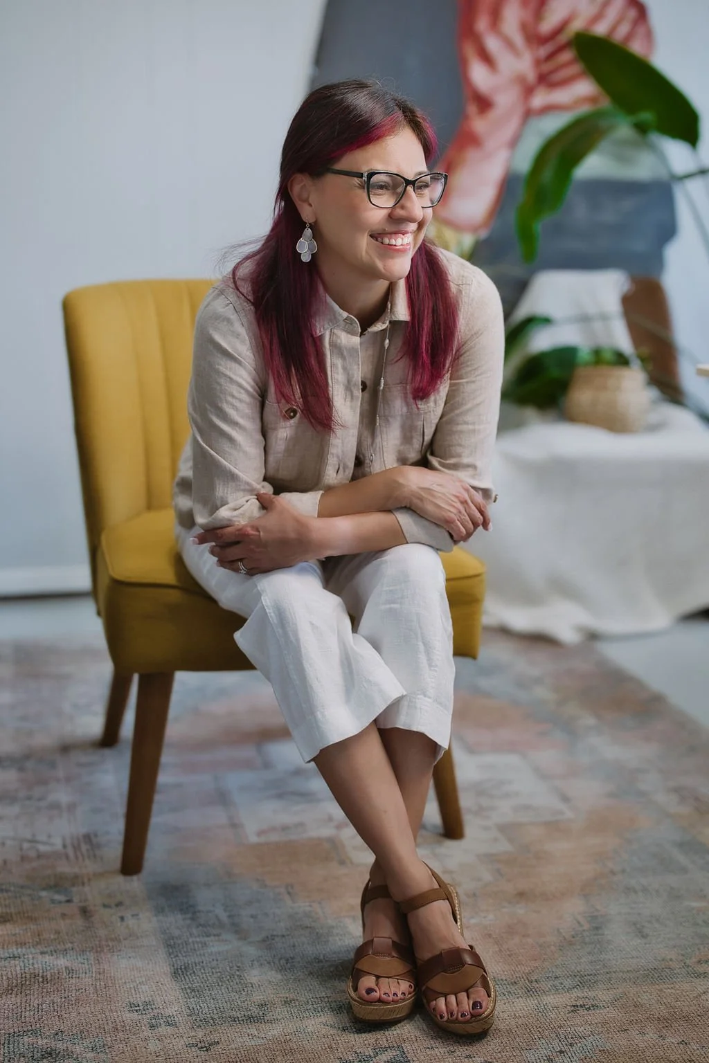 Veronica Diaz sitting on a yellow chair, smiling, in a room with plants and artwork in the background.