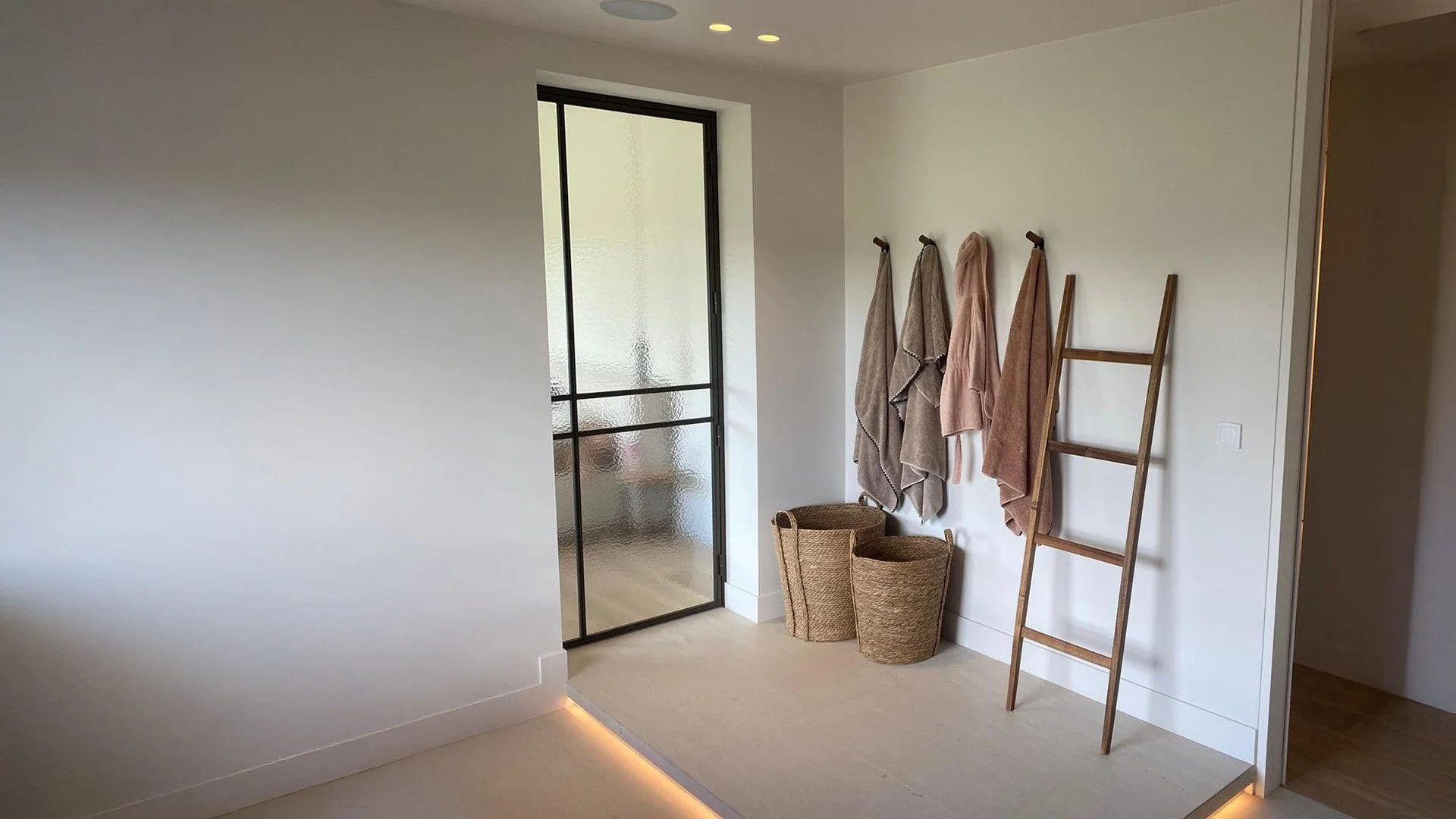 Een minimalistich ingerichte ruimte met handdoeken aan een muur, een ladder, twee manden en een glazen deur.