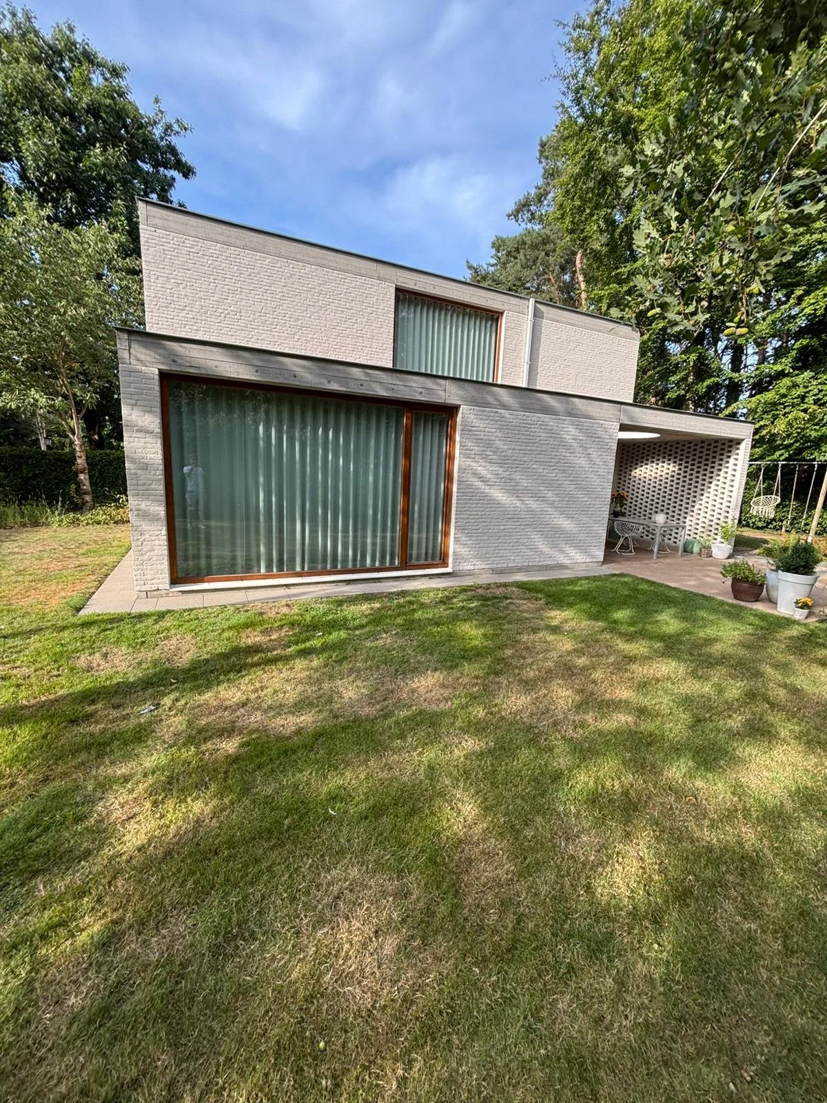 Moderne witte woning met grote ramen, terras en groene tuin in de buitenlucht onder een blauwe hemel.