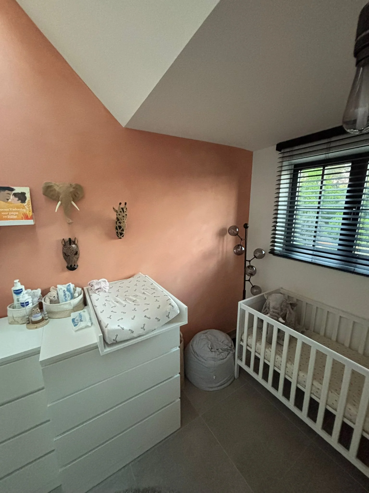 Babykamer met wit ledikant, muur met dierenvacht en dierenhoofd decoratie, commode met babydoekjes en verzorgingsproducten, venster met zwarte jaloezieën.