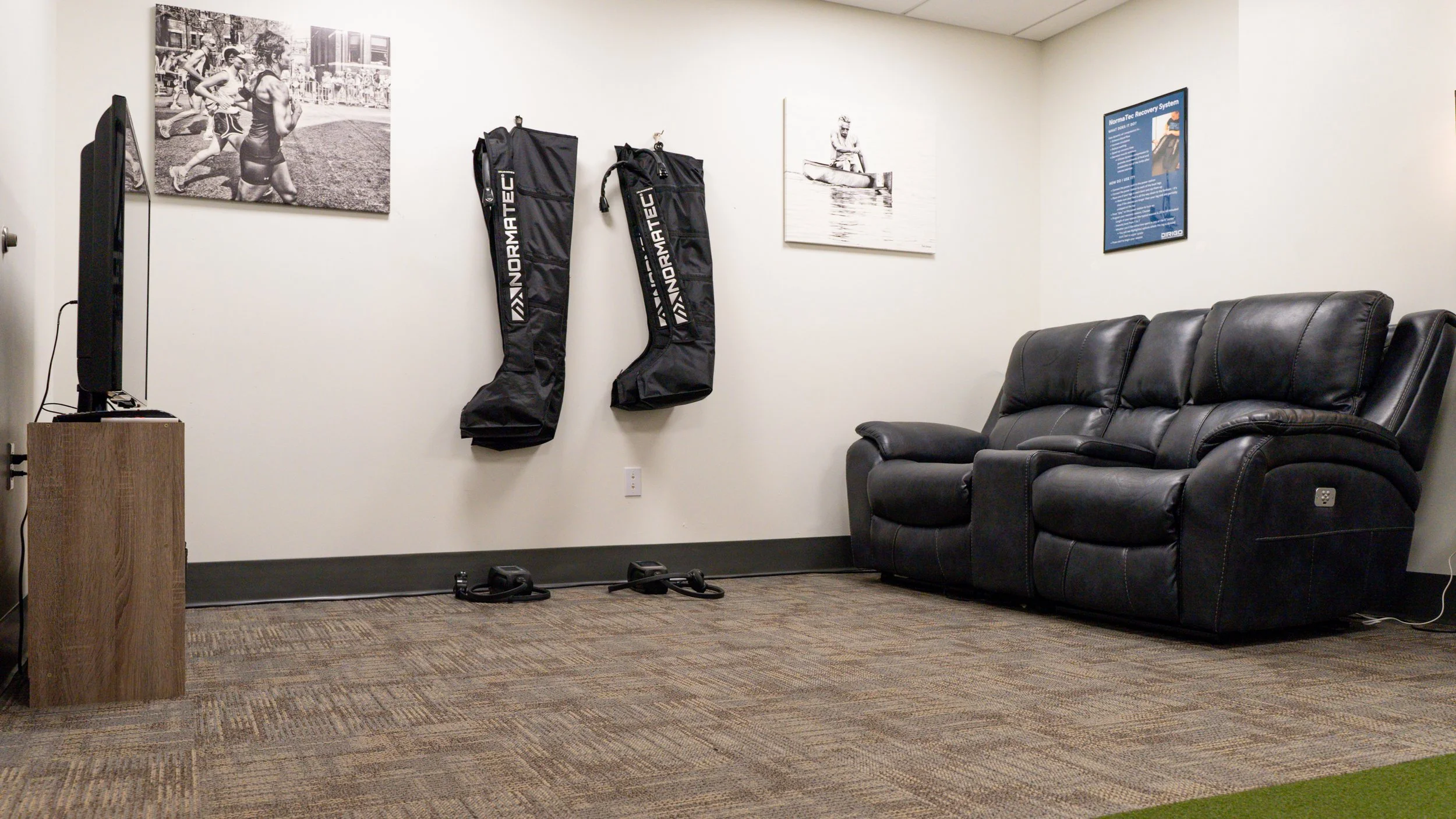 Room with a black recliner sofa, wall-mounted compression therapy boots labeled "Normatec," a TV on a wooden stand, and sports-themed wall art.