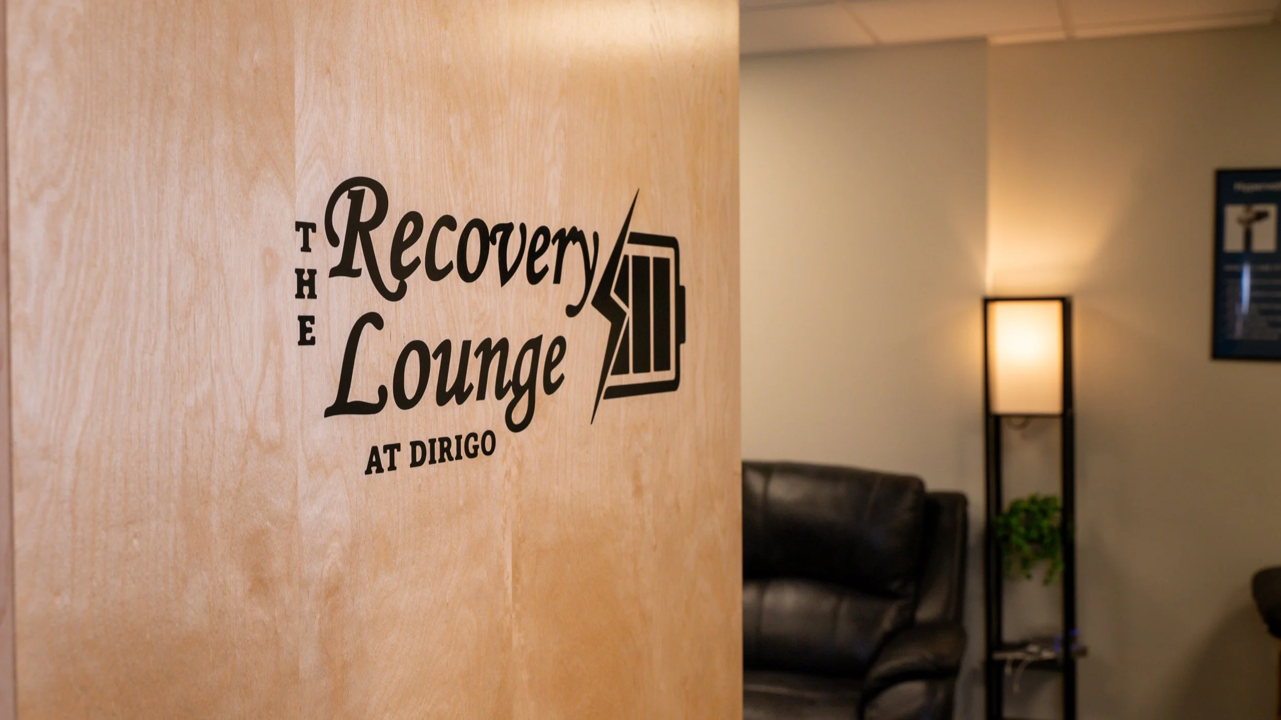 Sign reading "The Recovery Lounge at Dirigo" on a wood panel, with a leather chair and tall lamp in the background of a cozy room.