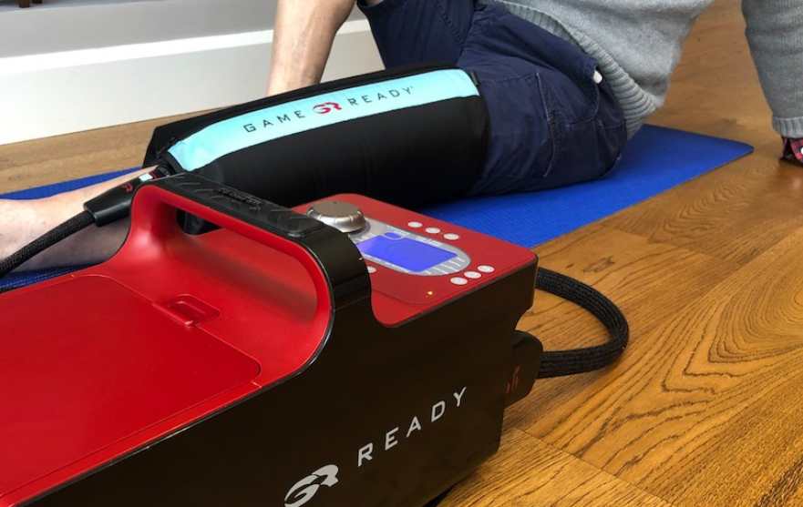 Person using Game Ready system on knee with control unit and wrap on wooden floor.