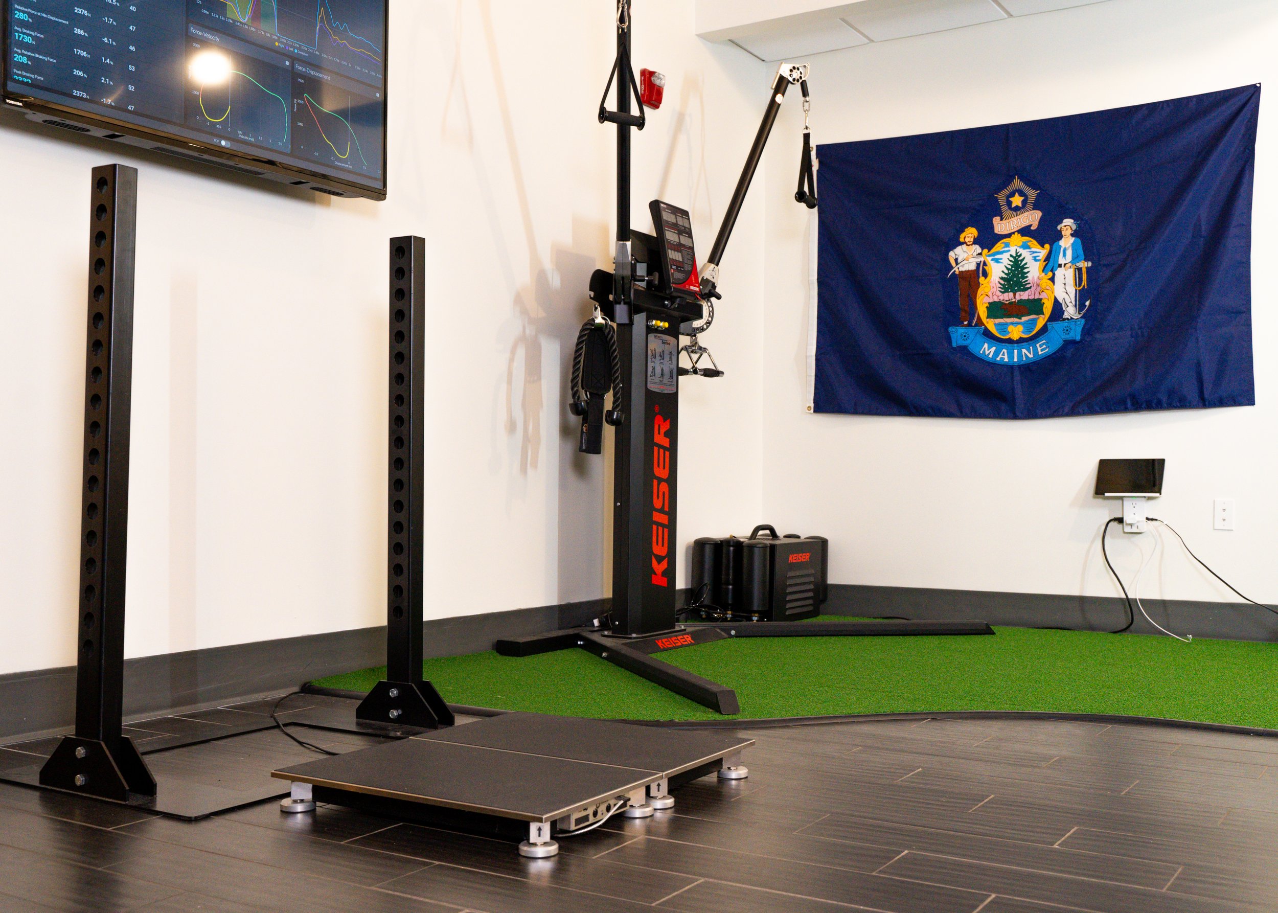 A gym setup featuring a Keiser functional trainer, two upright supports, a balance platform, and a Maine state flag on the wall. There is a screen displaying data graphs in the background.