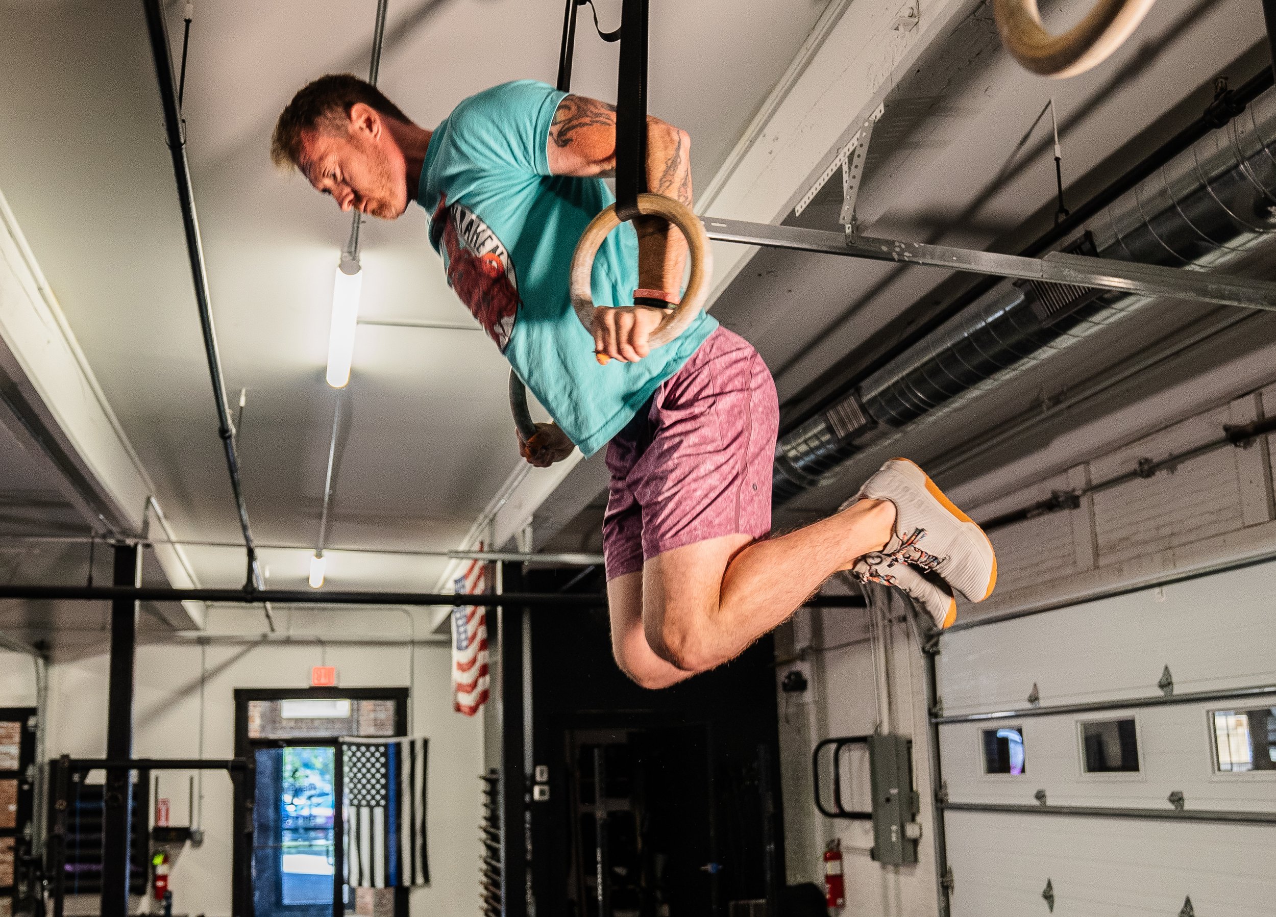A man performing a ring muscle-up in a gym, wearing a turquoise shirt, pink shorts, and white sneakers. The gym has an American flag and pipes on the ceiling.