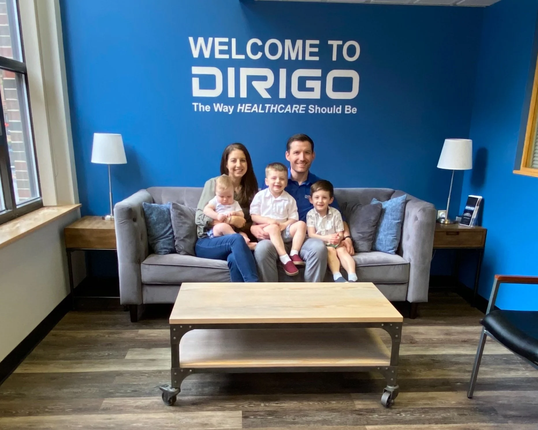 A family sitting on a gray sofa in an office lobby with blue wall displaying the text 'Welcome to DIRIGO The Way Healthcare Should Be.' The family includes two adults and three young children, and there are two lamps and wooden side tables beside the sofa.