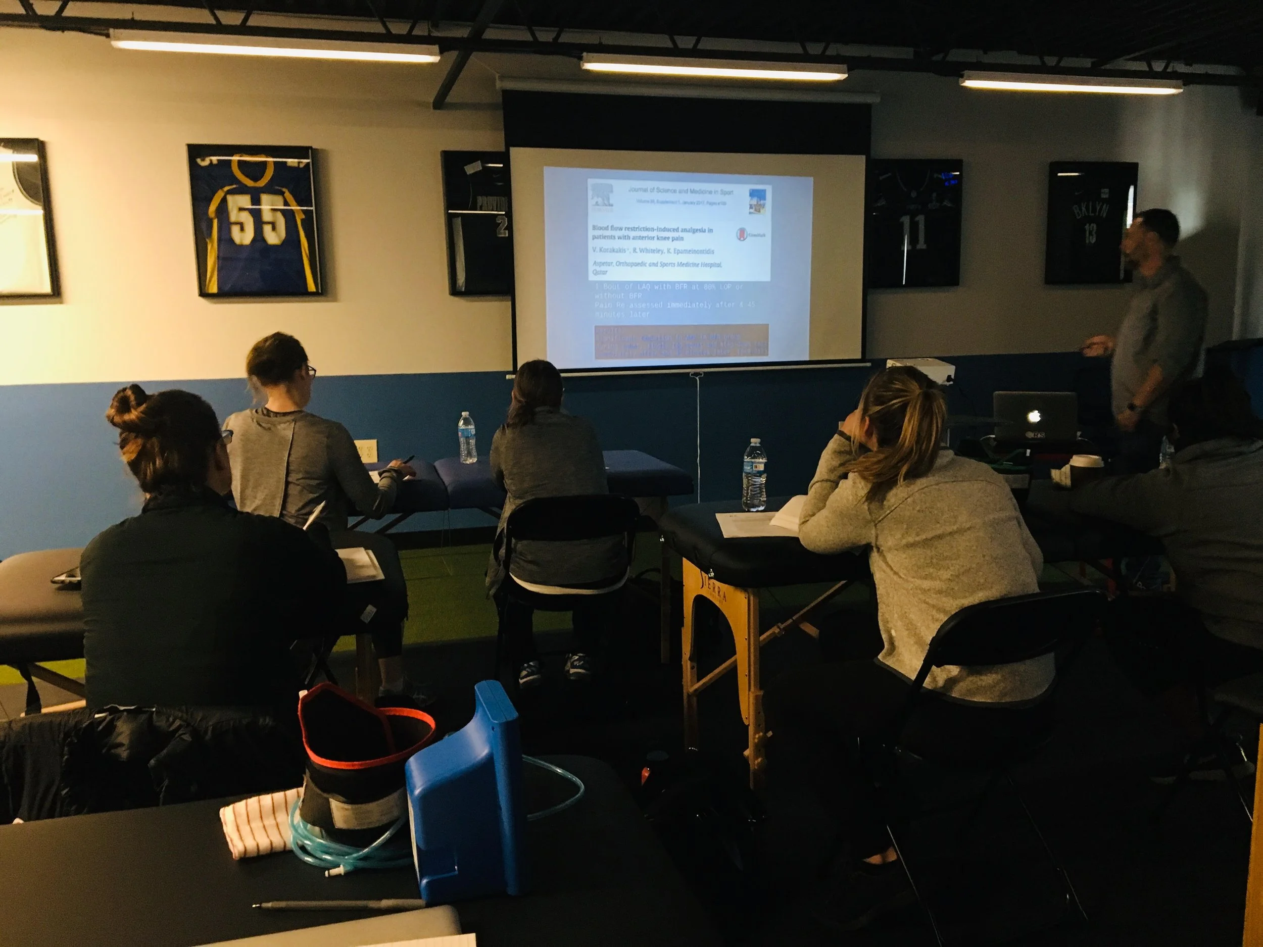 Classroom setting with students sitting at tables, facing a projector screen with a presentation on it. Sports jerseys are displayed on the wall.