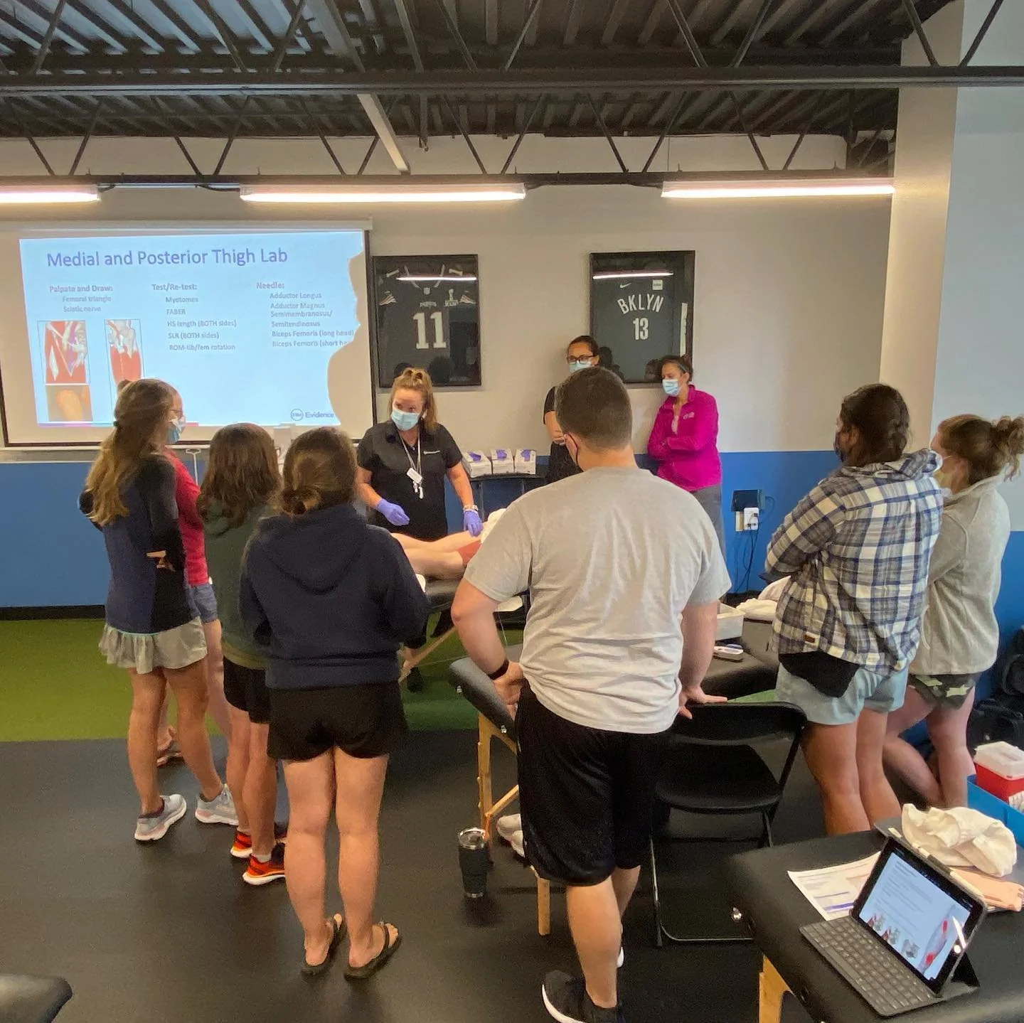 Group of people in a medical lab setting listening to an instructor near a presentation slide labeled 'Medial and Posterior Thigh Lab.'
