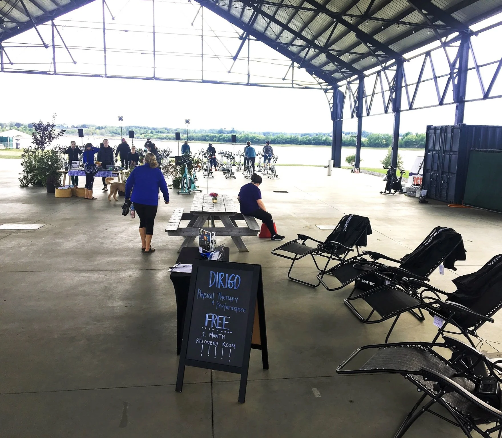 Outdoor activity space with exercise bikes, tables, and lounge chairs under a large open structure. A sign advertises free physical therapy sessions. People are gathered near the bikes, and the area overlooks a scenic view of grass and trees.
