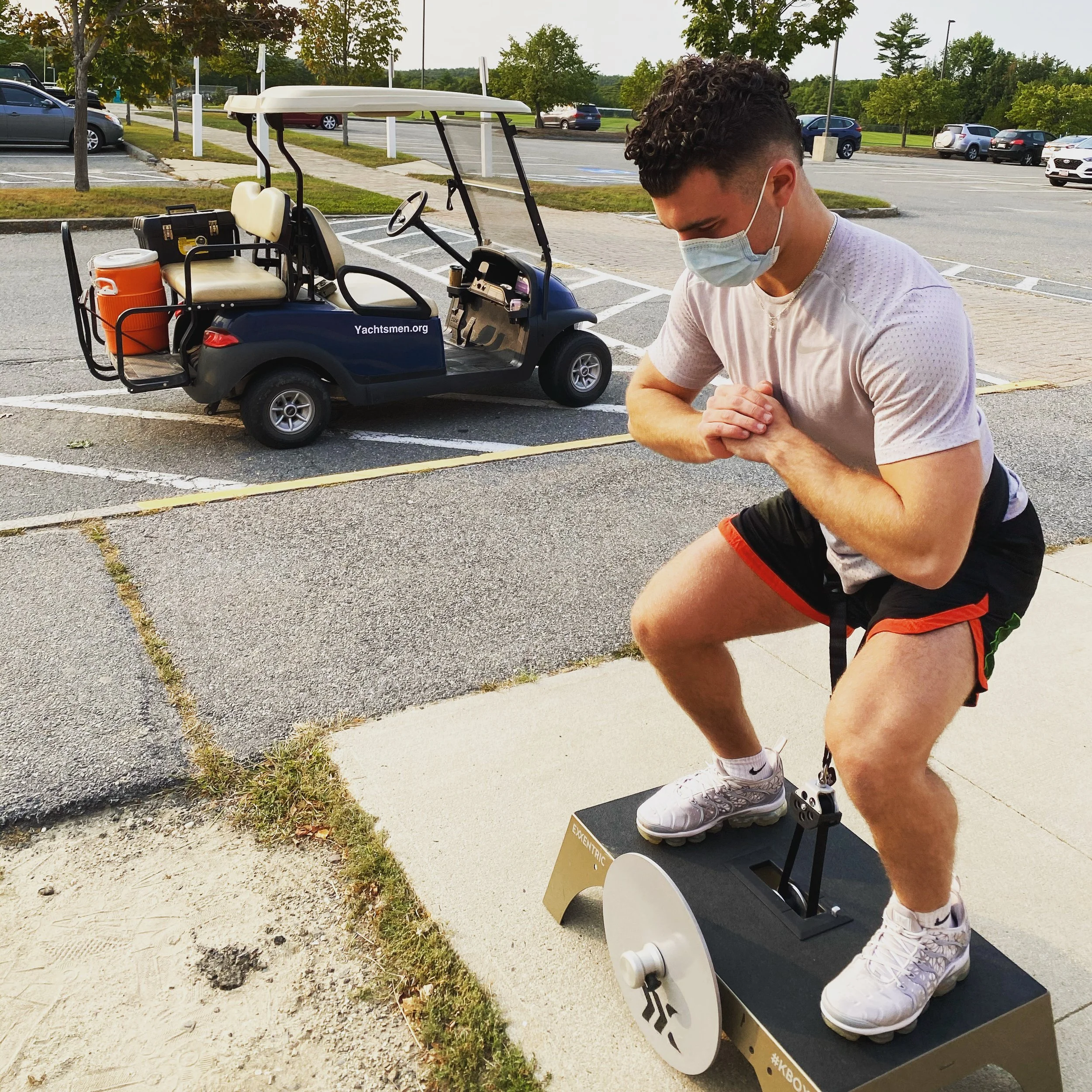 Man wearing a mask squatting on exercise equipment outside near a parked golf cart in an empty parking lot.