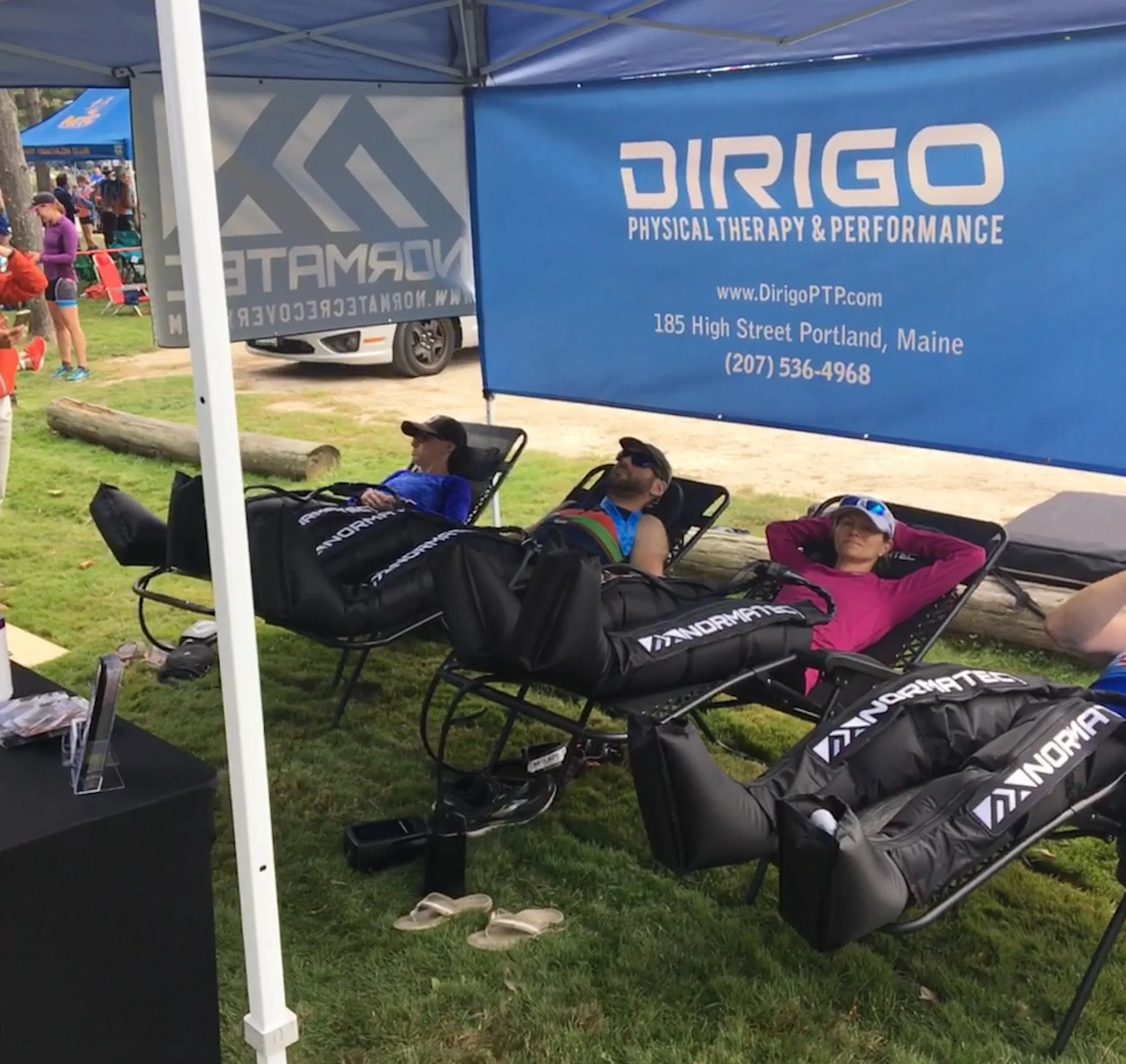 People using compression therapy devices at an outdoor event under a tent with a Dirigo Physical Therapy & Performance banner.