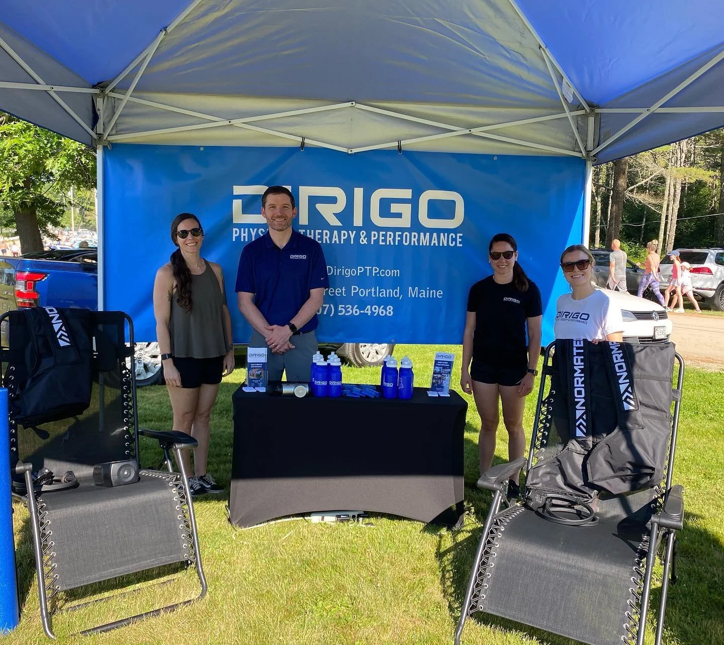 A group of four people standing under a canopy at an outdoor event. They are in front of a banner for Dirigo Physical Therapy and Performance. Two zero gravity chairs with Normatec branding are positioned on both sides of the table, which displays pr