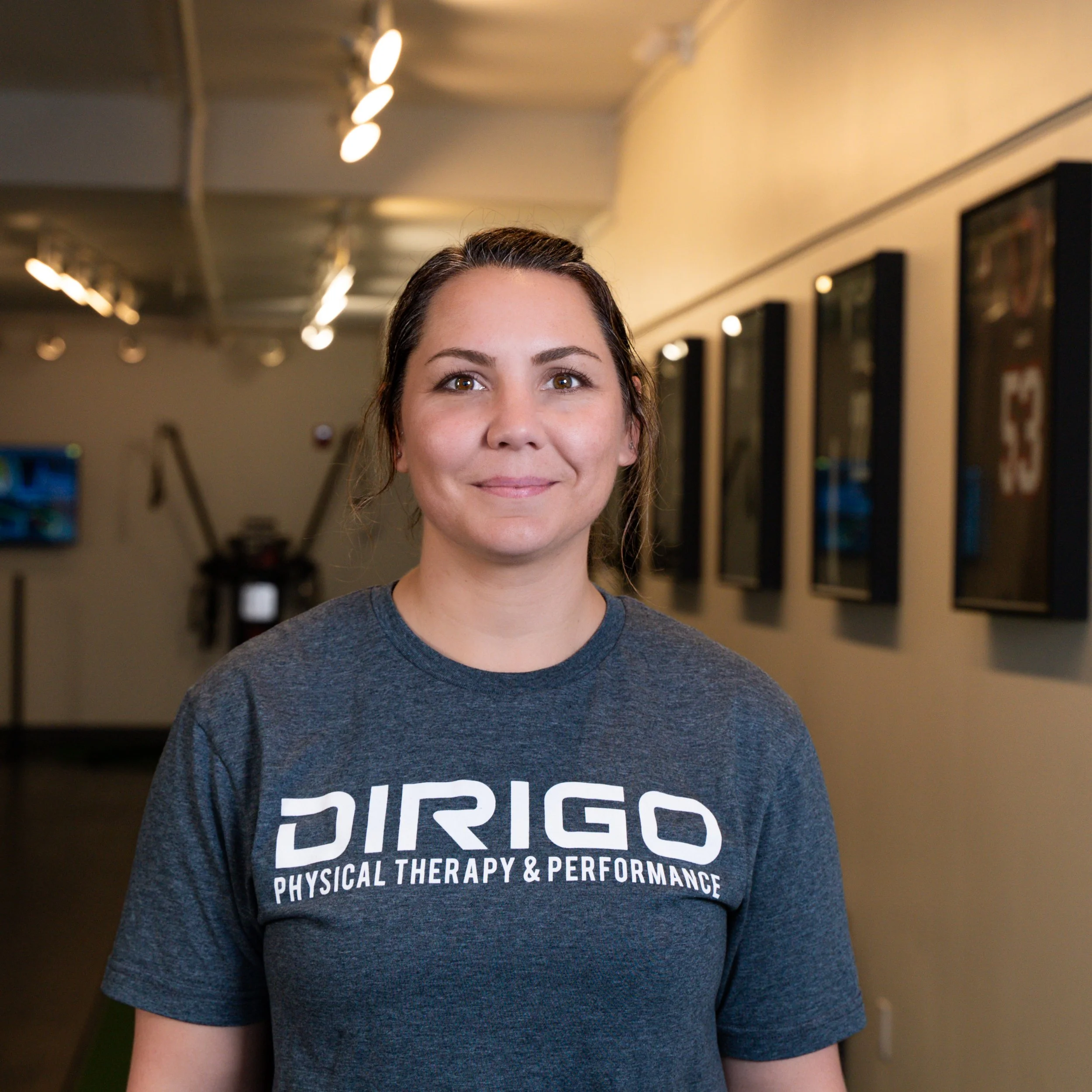Person in a "DIRIGO Physical Therapy & Performance" t-shirt standing in a hallway with sports memorabilia on the walls.
