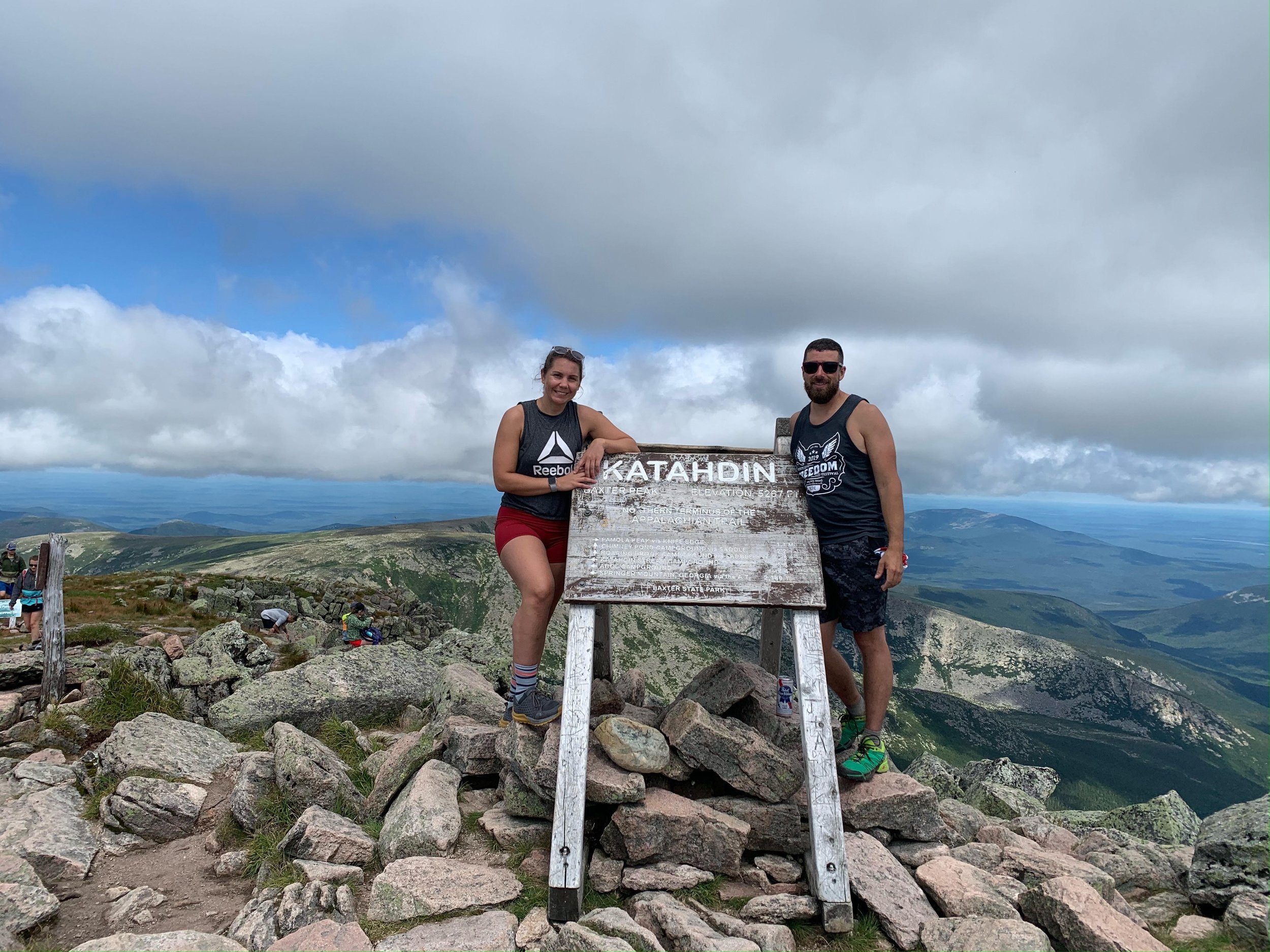 Two hikers standing on rocks next to a wooden sign at the summit of Mount Katahdin, surrounded by a mountainous landscape under a cloudy sky.