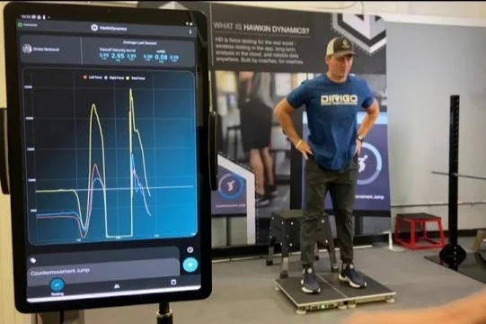 Man standing on sports performance equipment next to a screen displaying graphs and data, with a focus on physical metrics analysis.