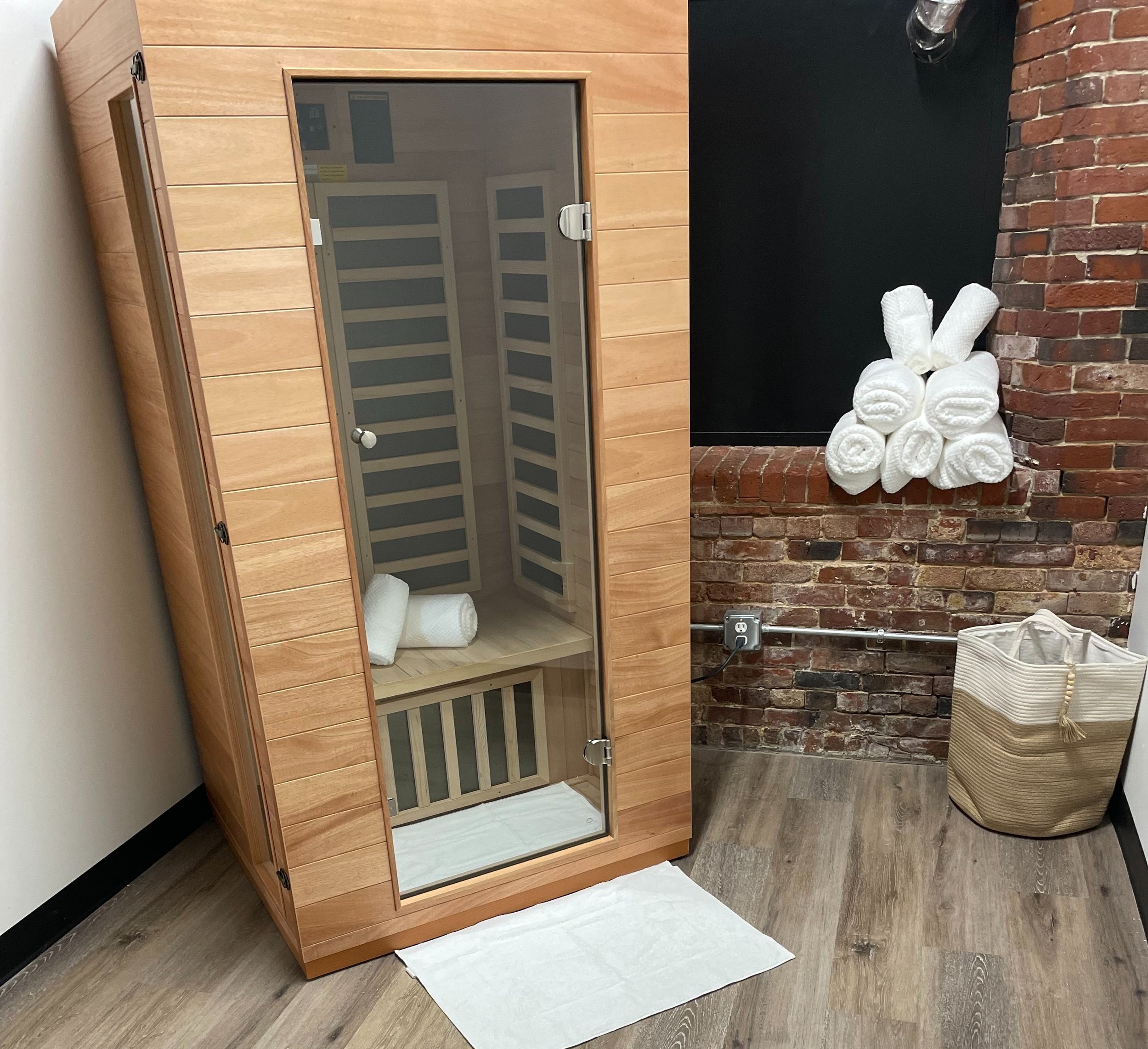 Wooden sauna room with a glass door, placed in a corner with light wood flooring and a brick wall. Inside are neatly stacked white towels. Additional rolled towels are stacked on a brick ledge, and a fabric basket is visible in the corner.