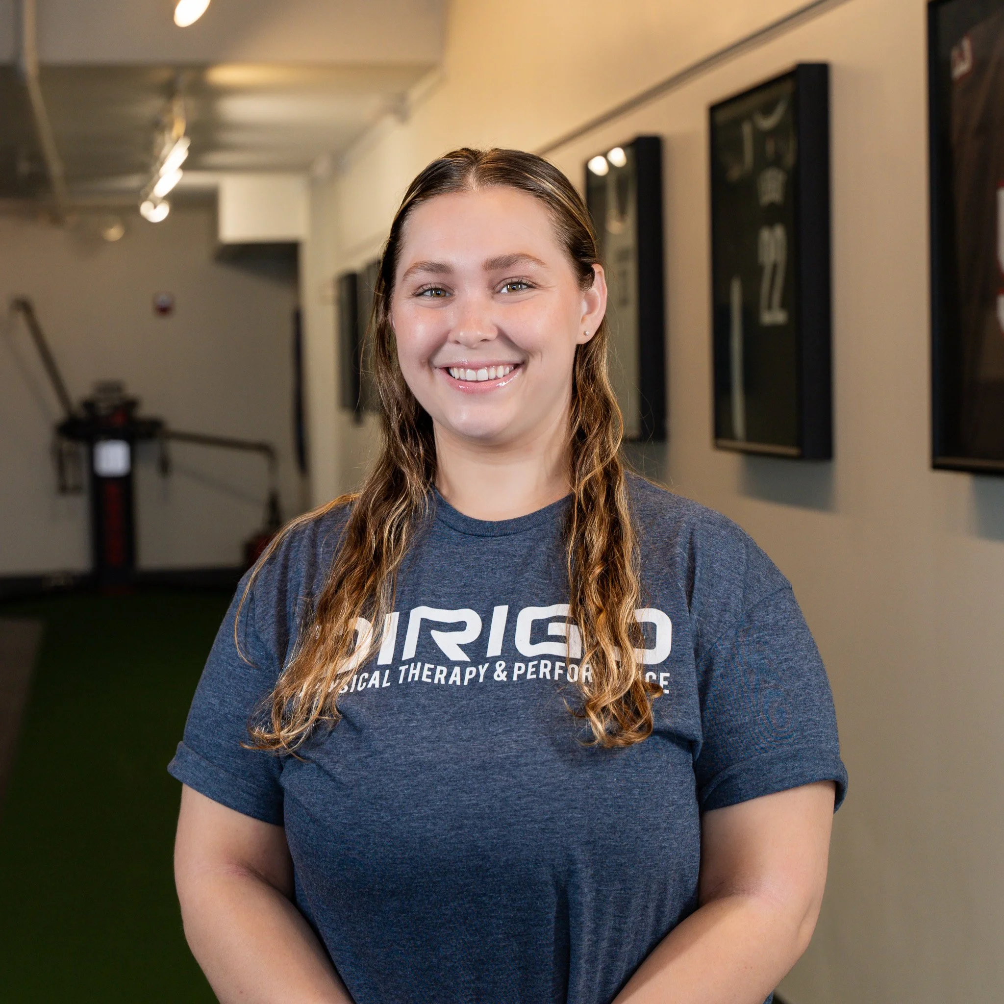 Woman smiling in a gym setting wearing a "Dirigo Physical Therapy & Performance" T-shirt.