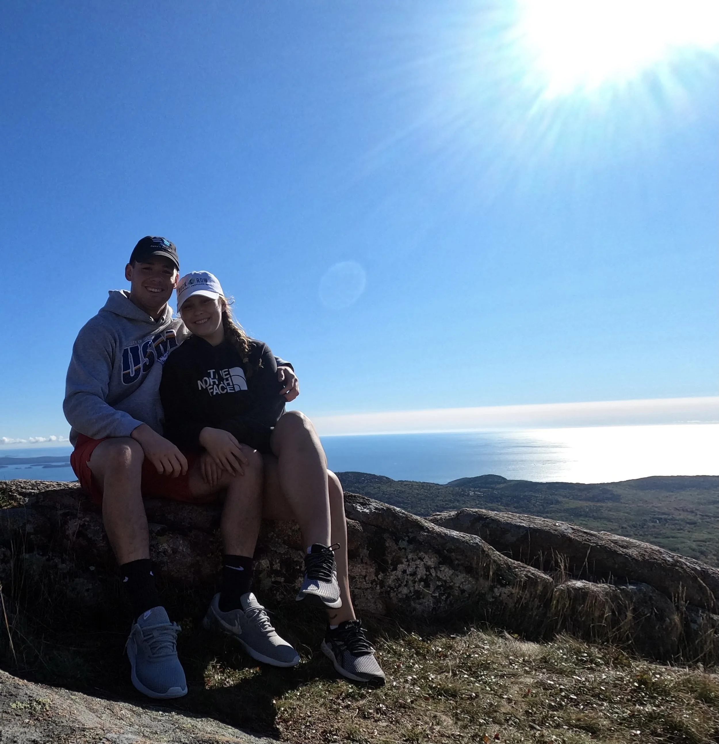 Couple sitting on a rocky hill with ocean view, wearing casual sportswear, under a clear blue sky with the sun shining brightly.
