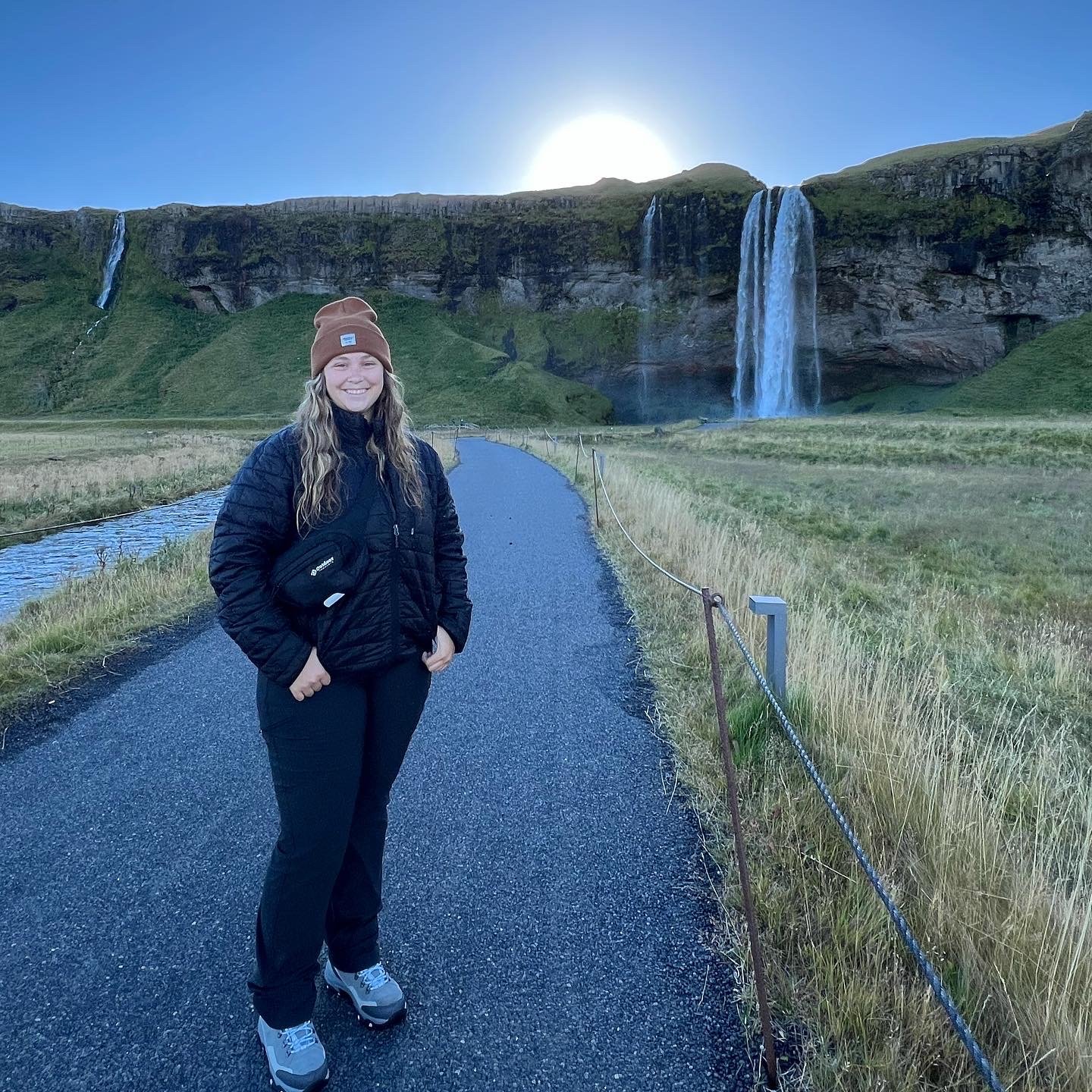 Woman smiling on a paved path with waterfalls and green cliffs in the background under a clear sky.