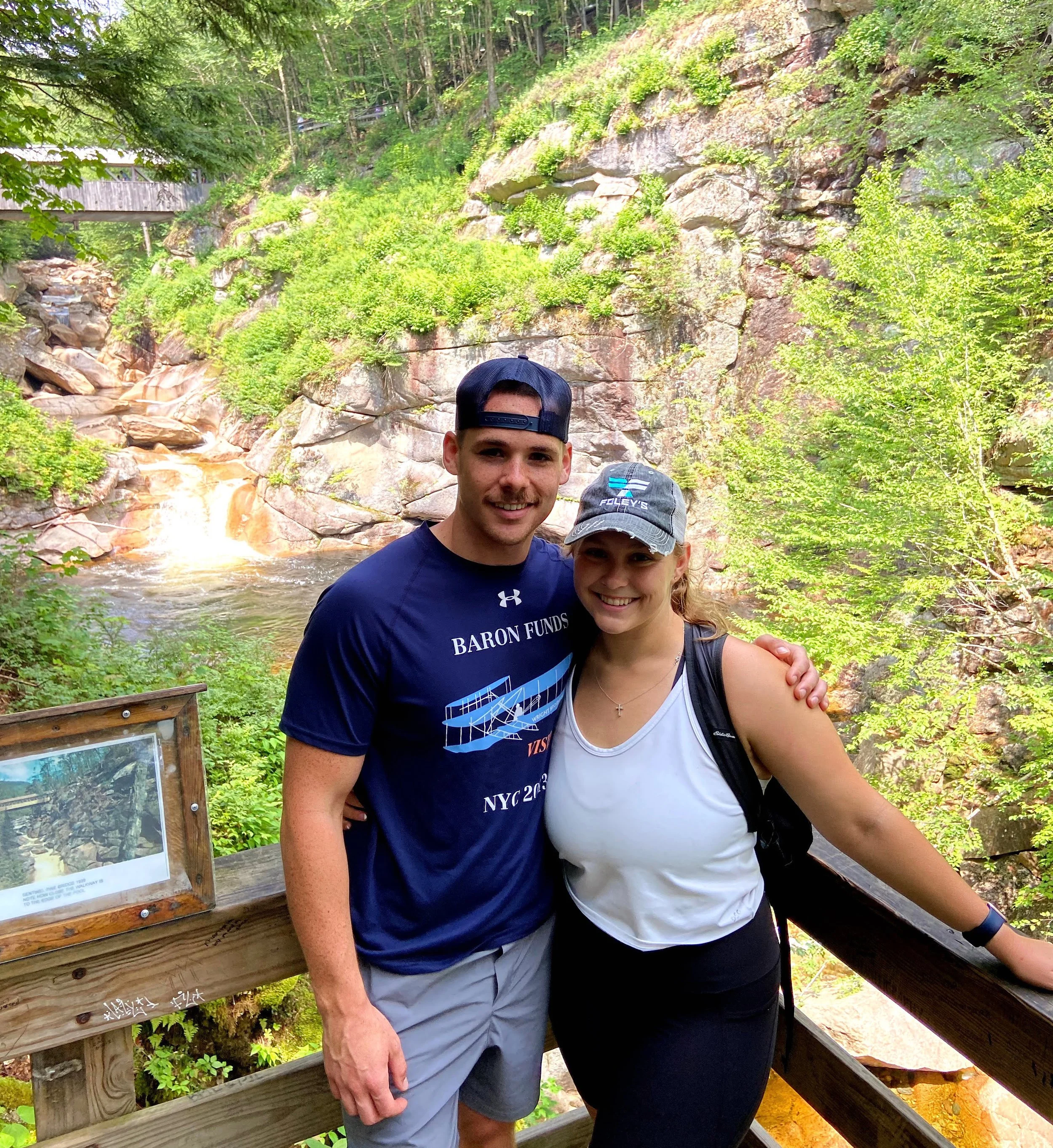 A couple standing on a wooden bridge in a scenic forest area with rocky cliffs and a waterfall in the background, surrounded by lush greenery.