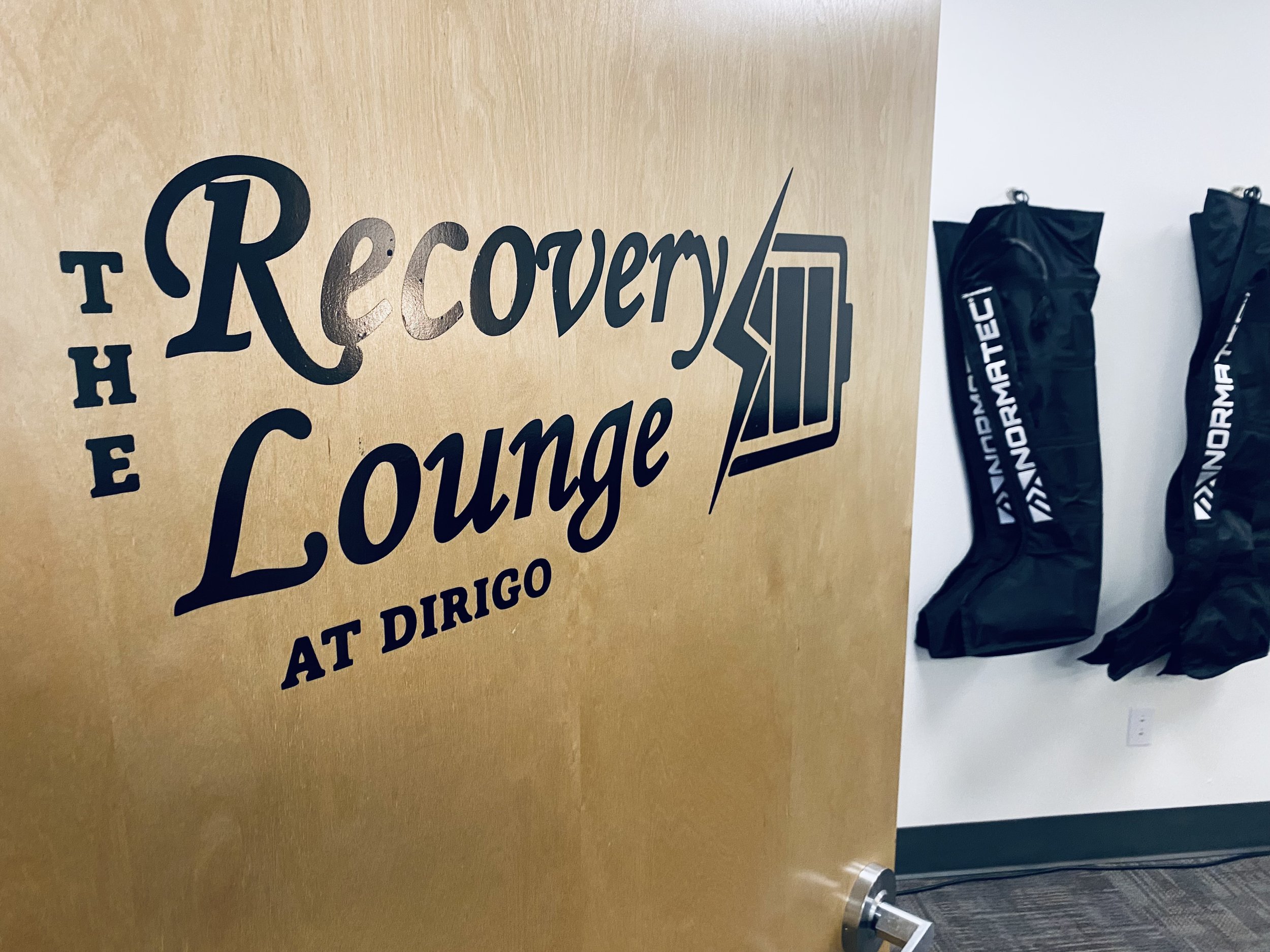 Door with text "The Recovery Lounge at Dirigo" and two Normatec recovery boots hanging on a wall.
