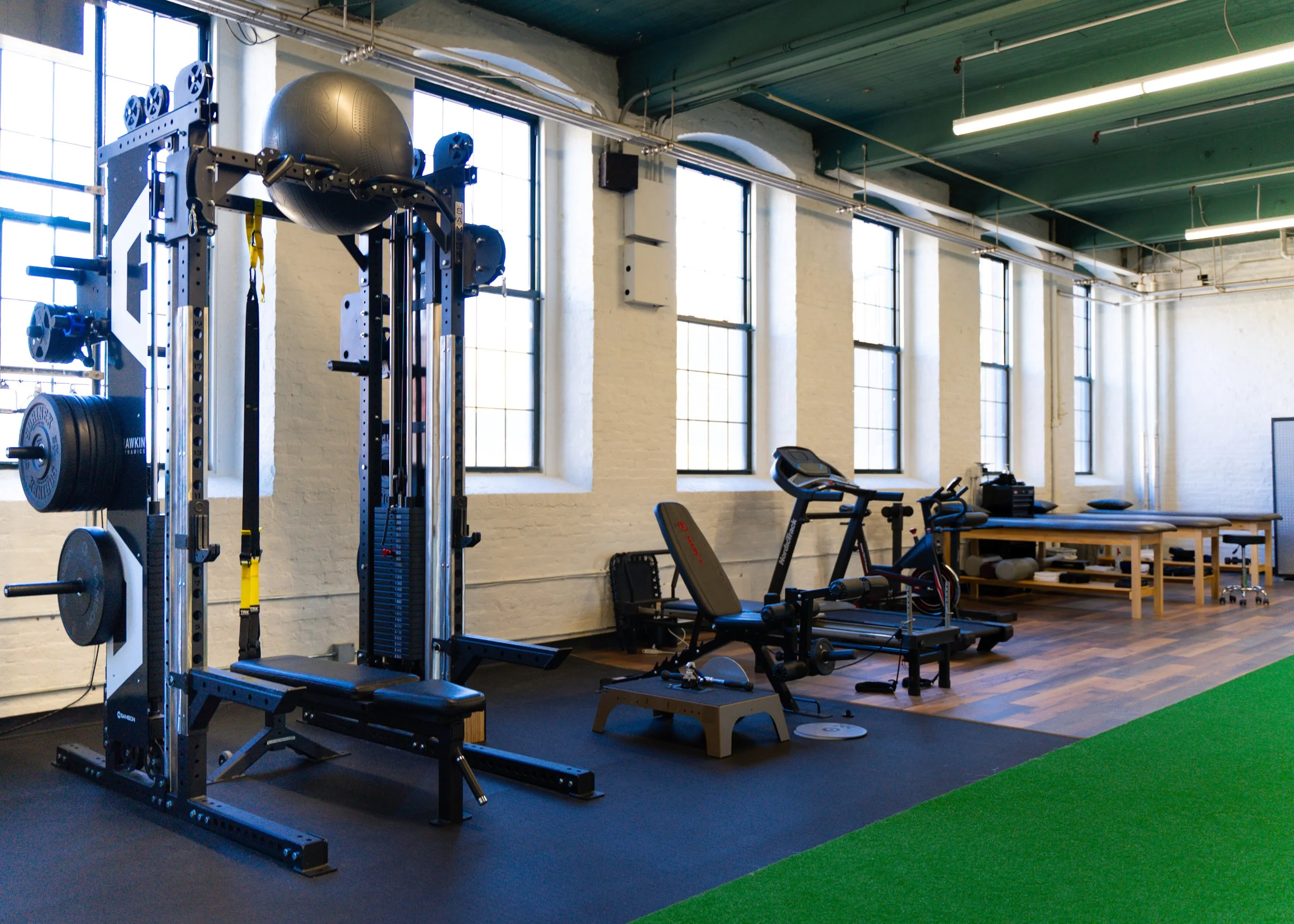 Gym with weightlifting equipment, treadmill, and exercise benches near large windows.