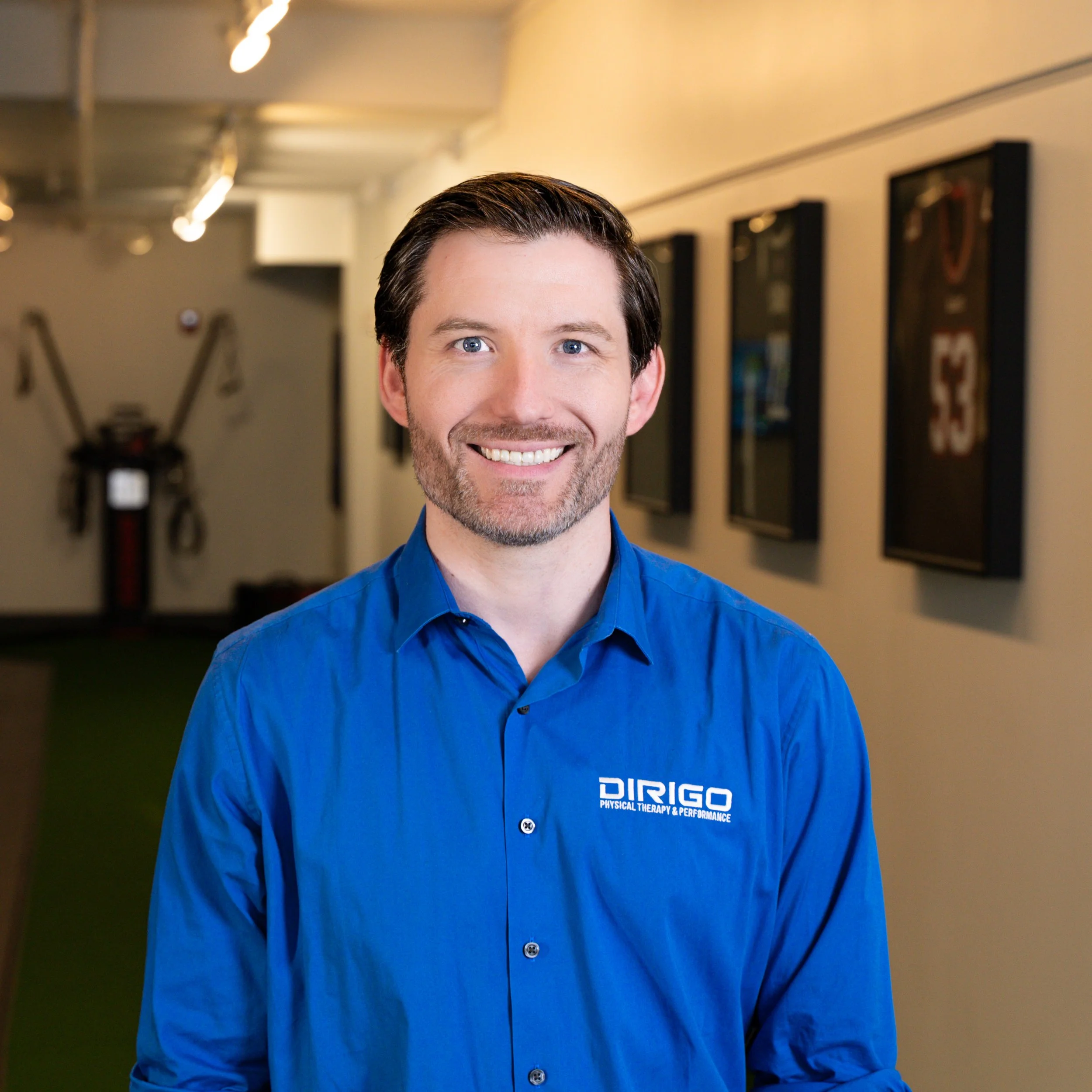 Man wearing a blue shirt with 'Dirigo Physical Therapy & Performance' logo stands in a hallway, smiling. Sports jerseys are displayed on the wall behind him. The setting appears to be a professional office or clinic with exercise equipment in the background.