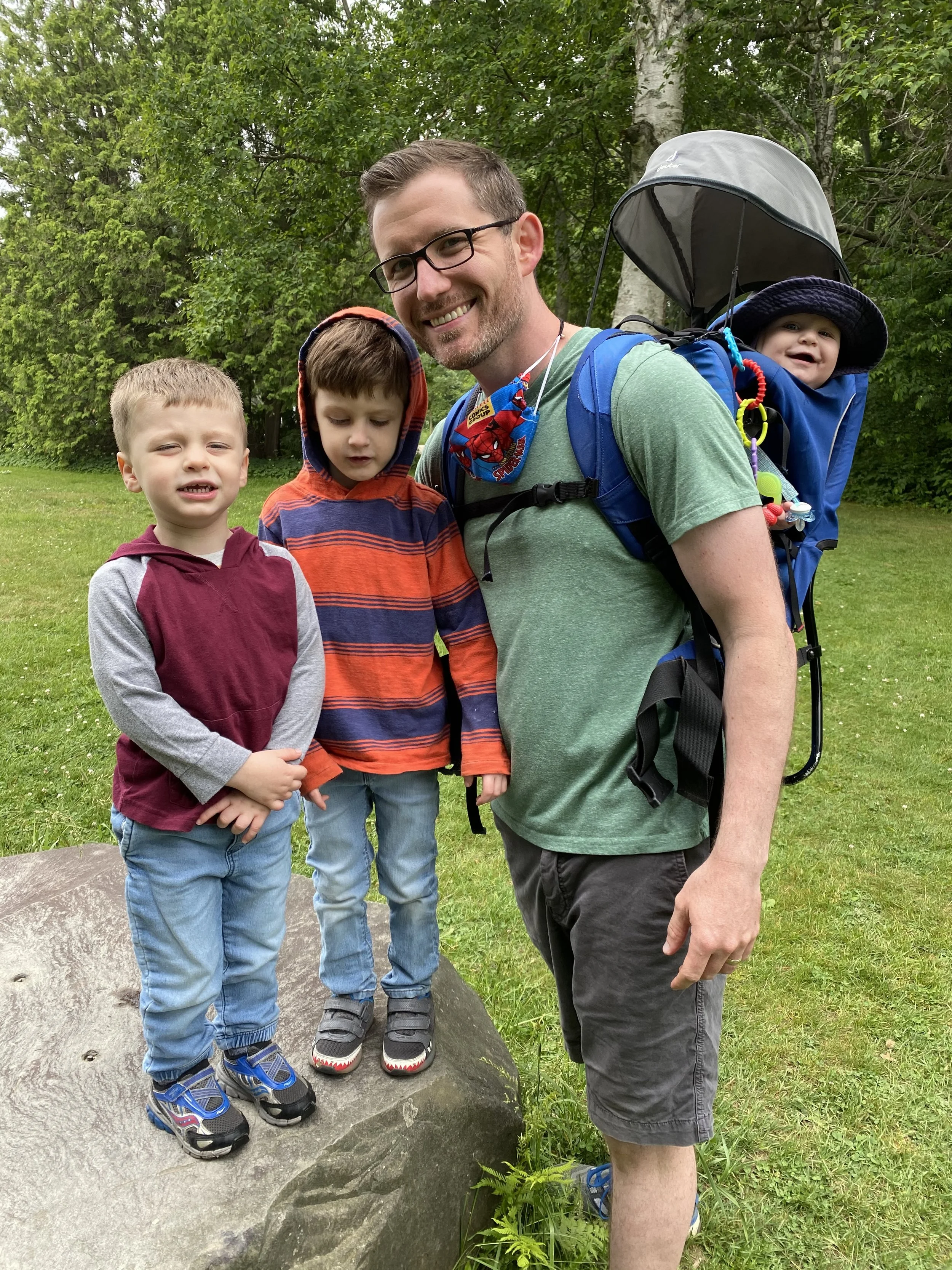 A smiling man wearing glasses and a green shirt carries a baby in a backpack carrier. Two young boys stand next to him on a large rock. The scene is set outdoors with a background of grass and trees.