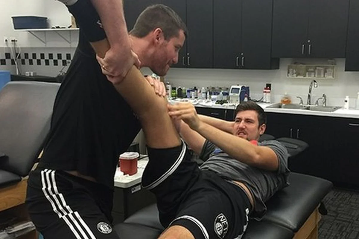 Two men in a physical therapy session: one man stretches the leg of another who is lying on a therapy bed. Both are wearing athletic clothing in a treatment room with a counter and sink in the background.