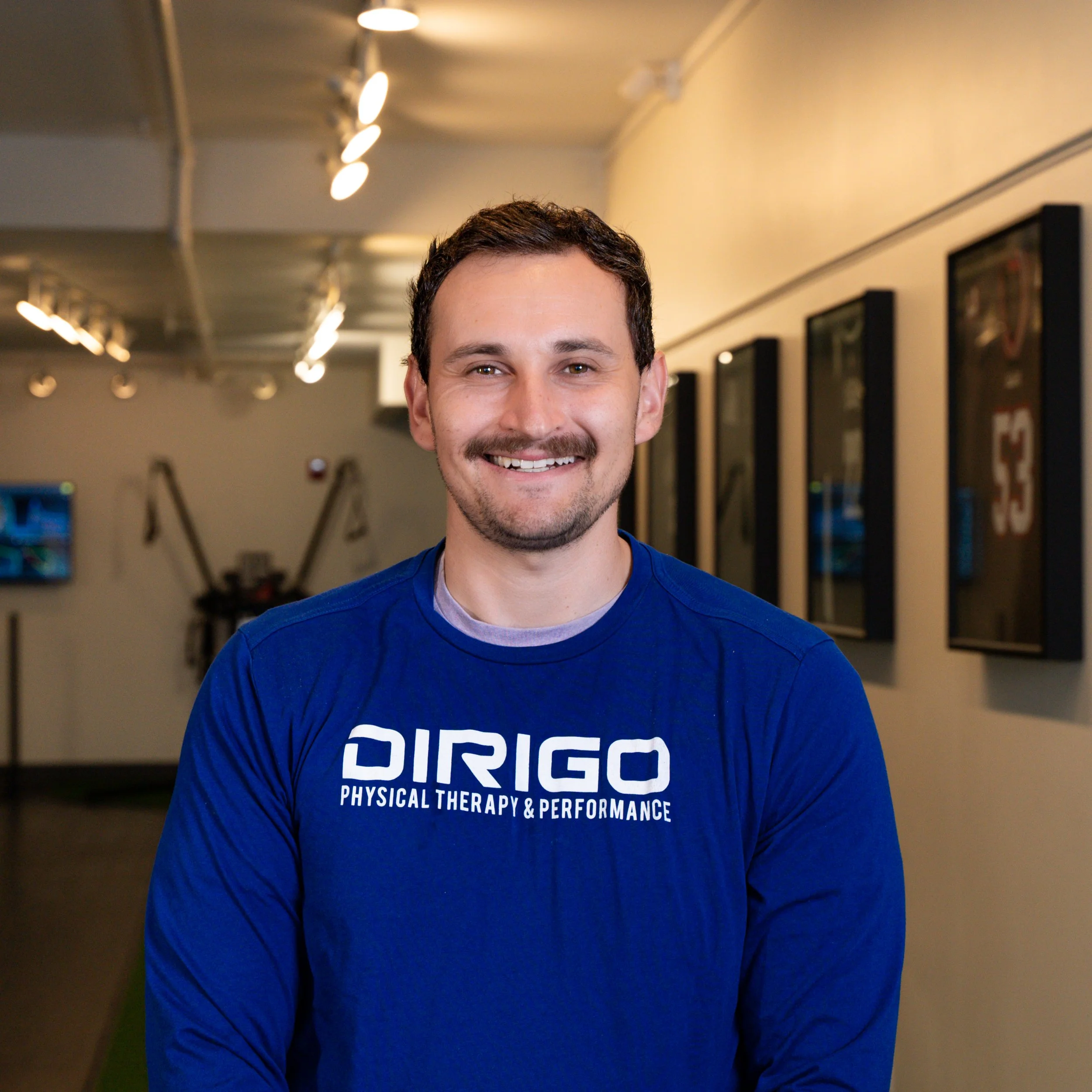 Man in a blue 'Dirigo Physical Therapy & Performance' shirt smiling in a sports facility.