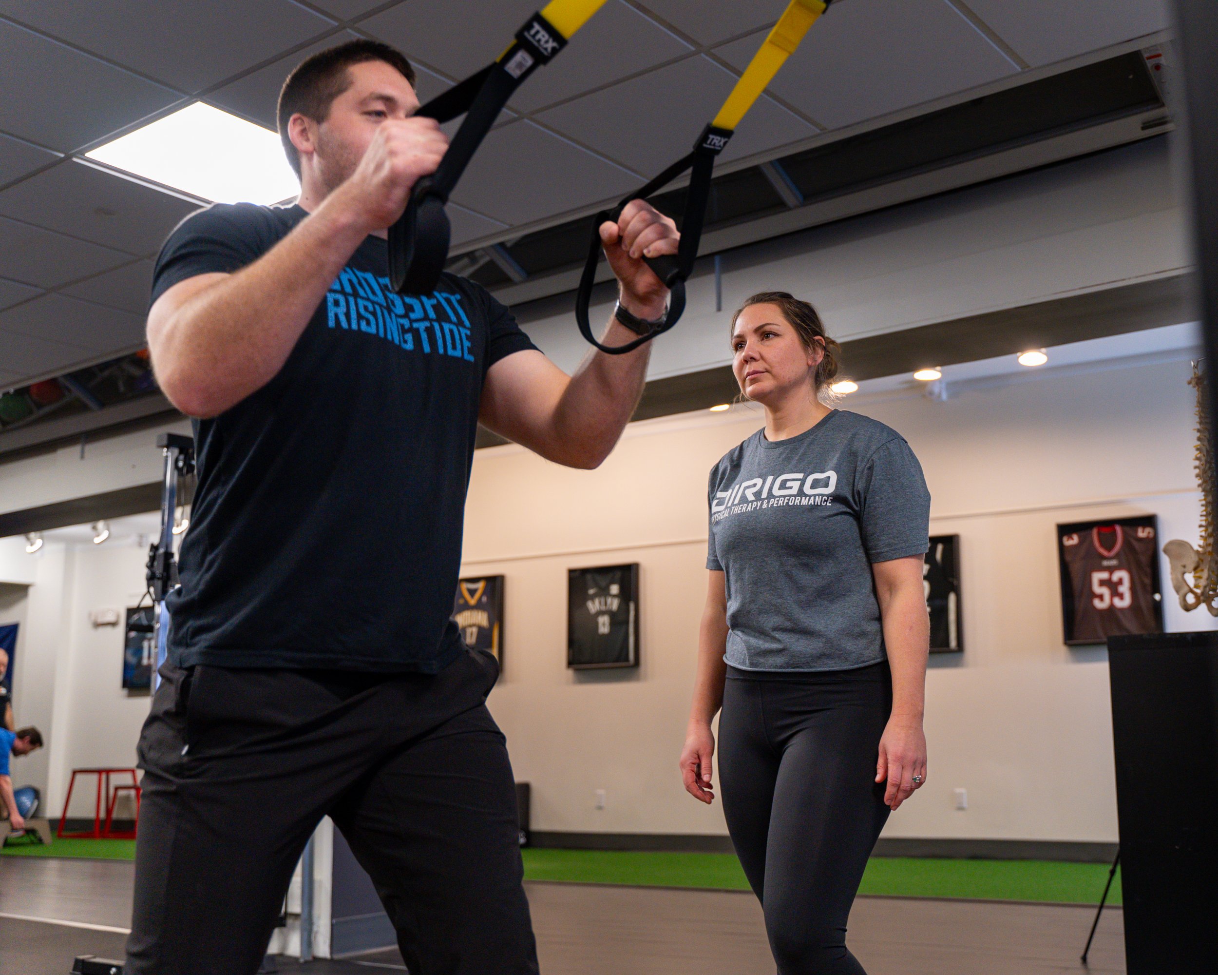 A man exercising with TRX straps while a woman observes in a gym setting. The gym has sports jerseys displayed on the walls.