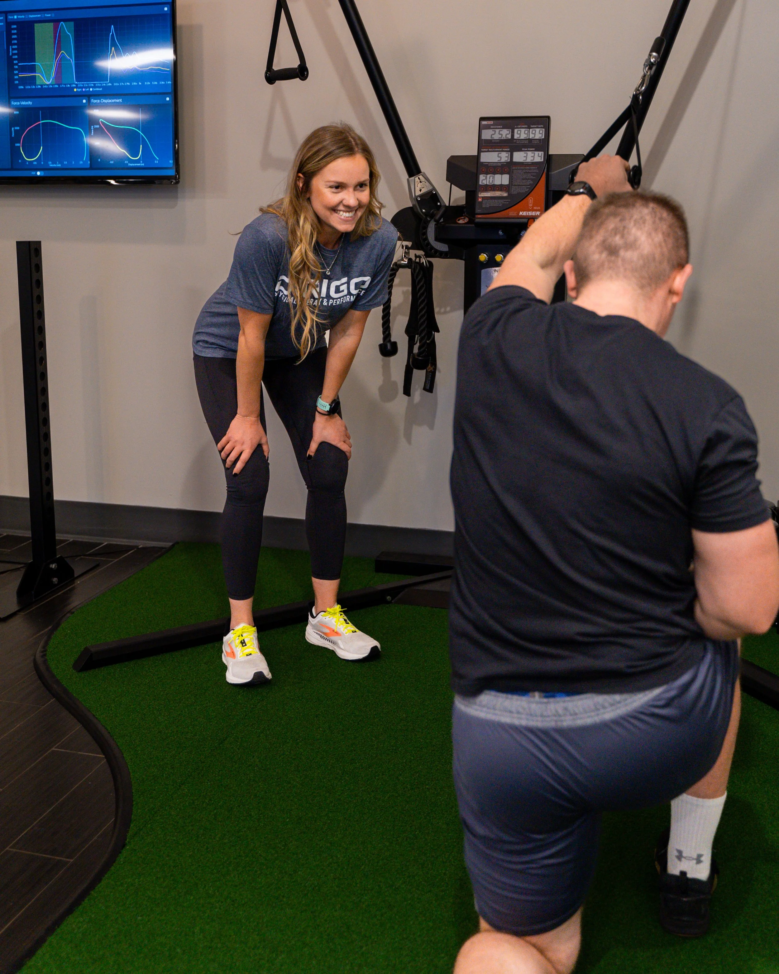 A woman in athletic apparel smiling at a man exercising indoors on artificial turf, with fitness equipment and a screen displaying data in the background.