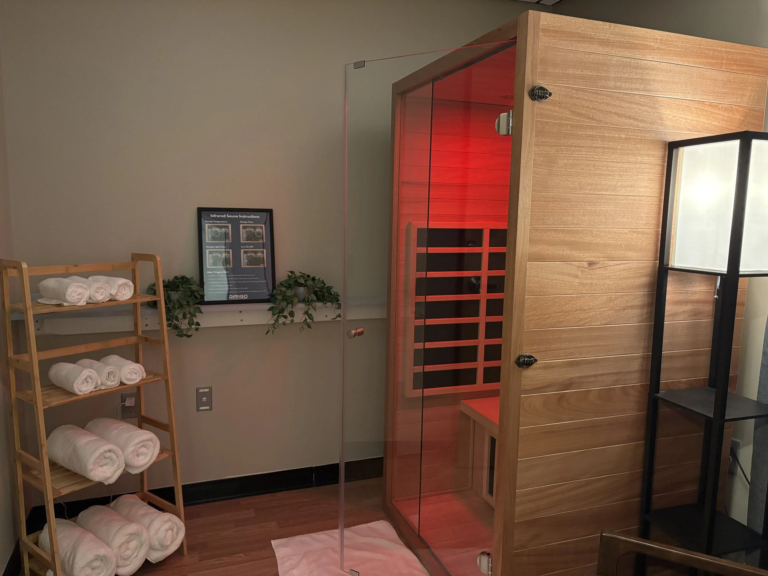 A small infrared sauna room with a wooden door, adjacent to a bamboo shelf holding rolled white towels and a nearby lamp.