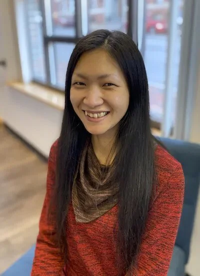 A person with long black hair and a red sweater is sitting in a room with large windows and wooden floors, smiling at the camera.