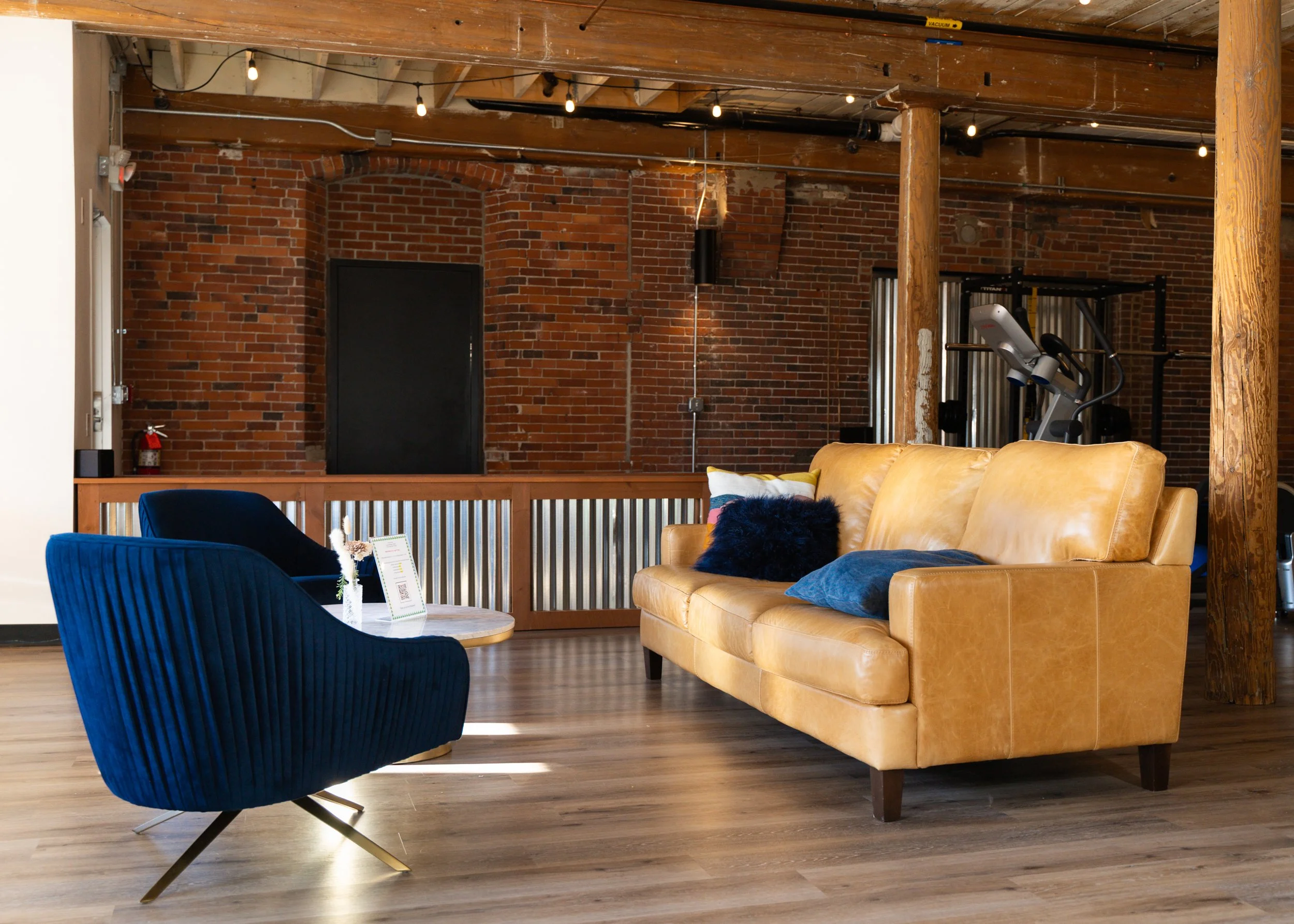Modern industrial living room with yellow leather couch, blue chairs, brick wall, and wooden beams.