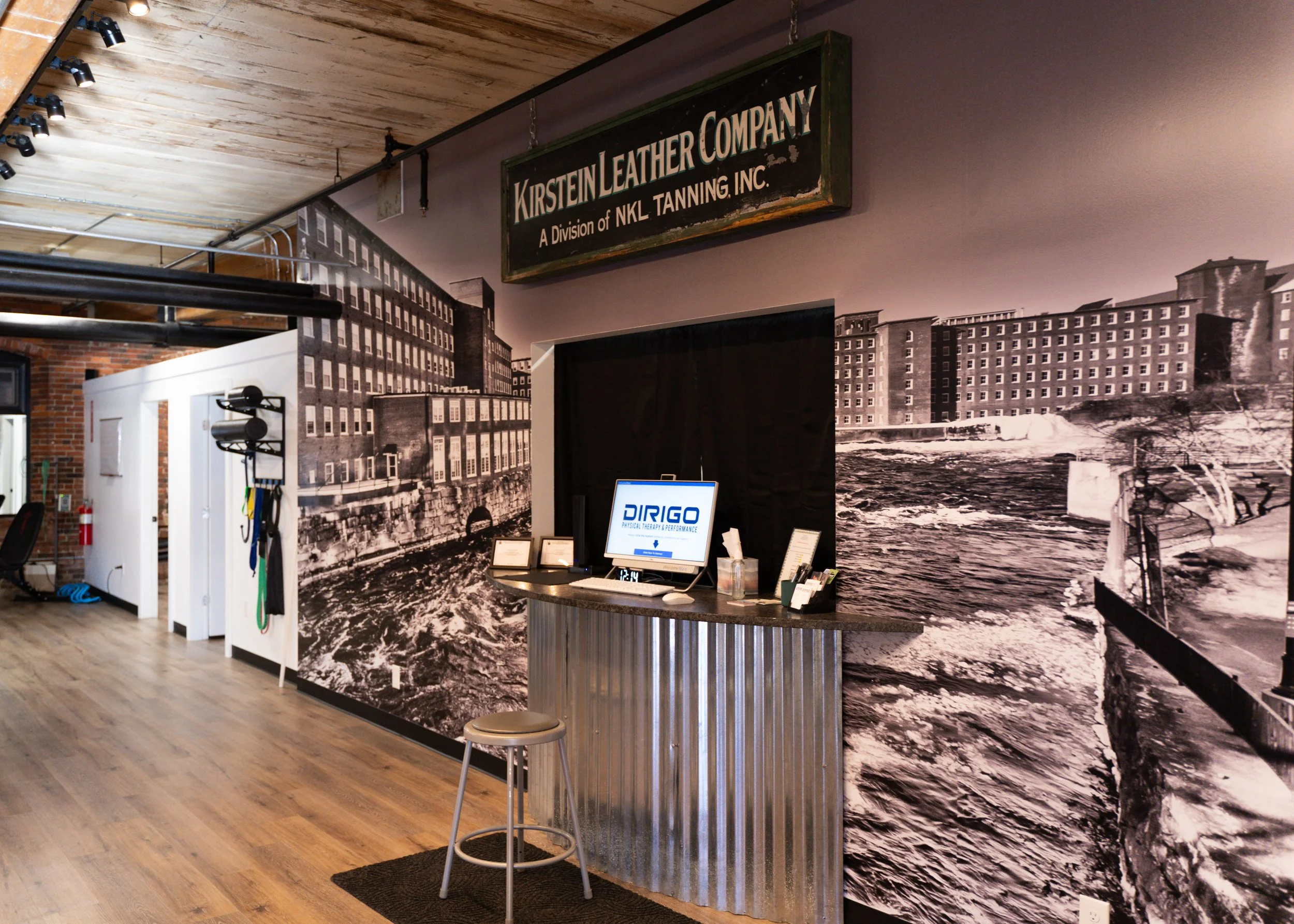 Interior of a commercial space with wooden flooring and industrial decor. A wall displays a large black-and-white historical photograph of a building with water. Above the entrance is a vintage sign for "Kirstein Leather Company." A small counter with a computer screen showing "DIRIGO" is near the front.