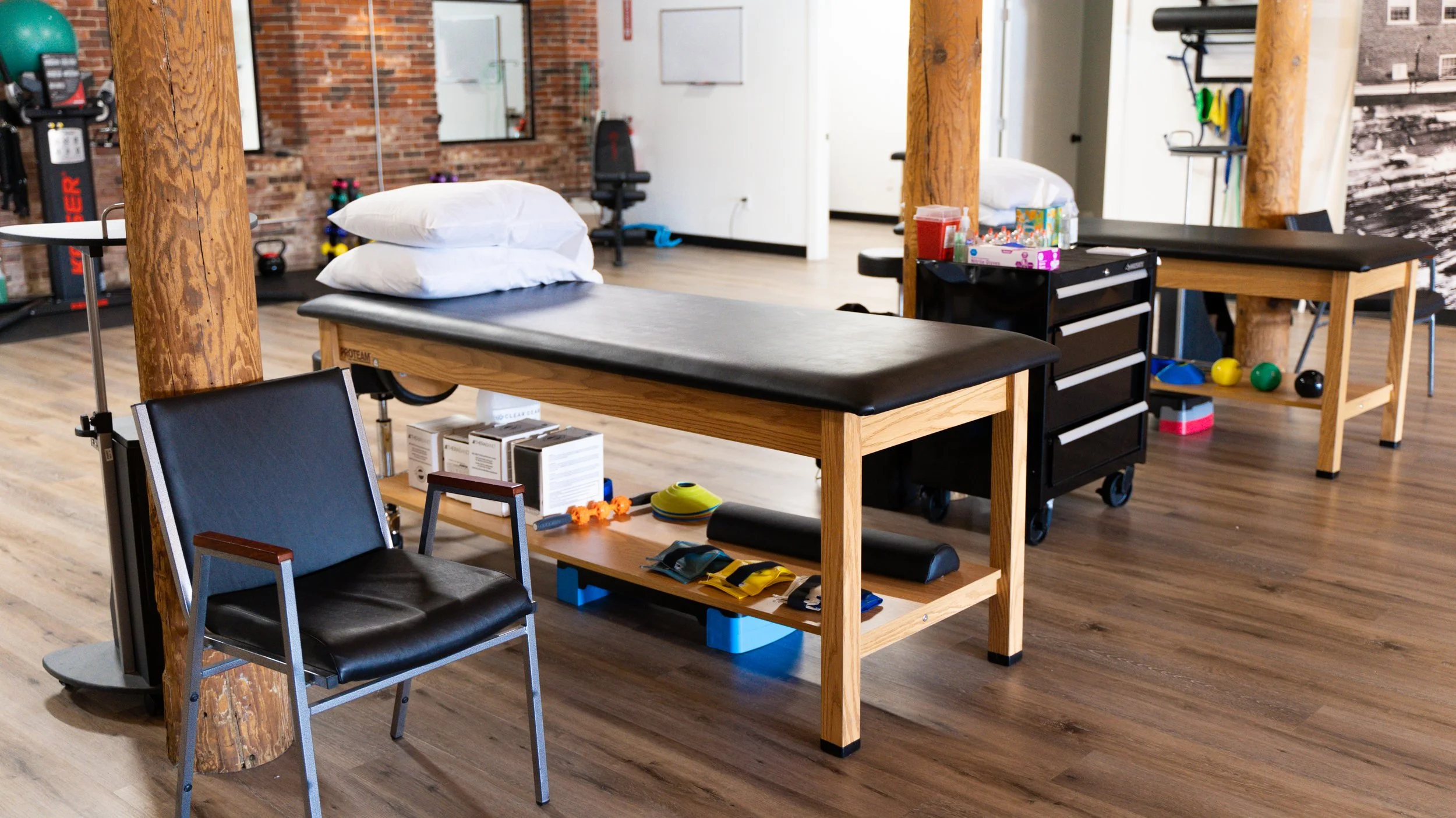 Physical therapy room with equipment, including an examination table, chairs, exercise tools, and storage cart, set against a wooden floor and brick wall backdrop.