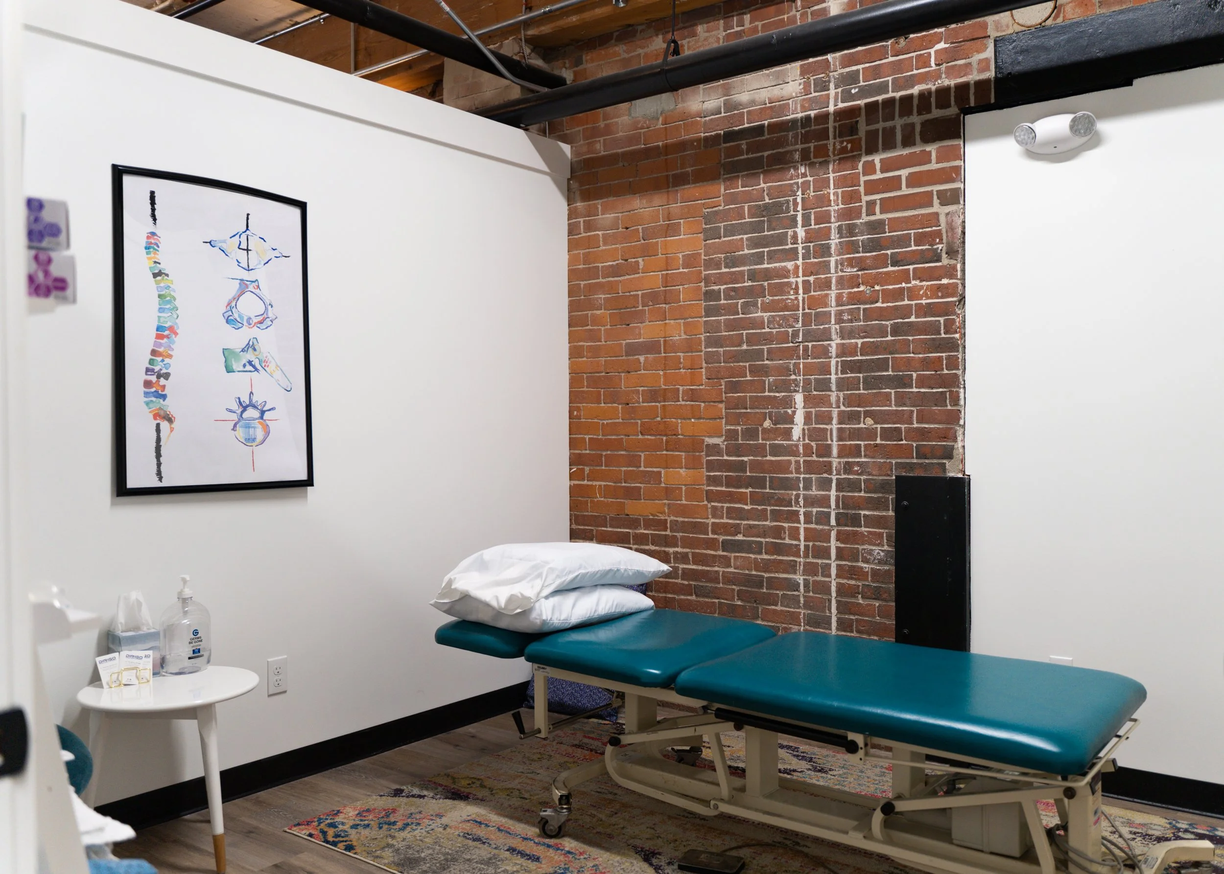 Chiropractic office with a massage table, skeletal anatomy chart, and a small side table with hand sanitizer and tissues.