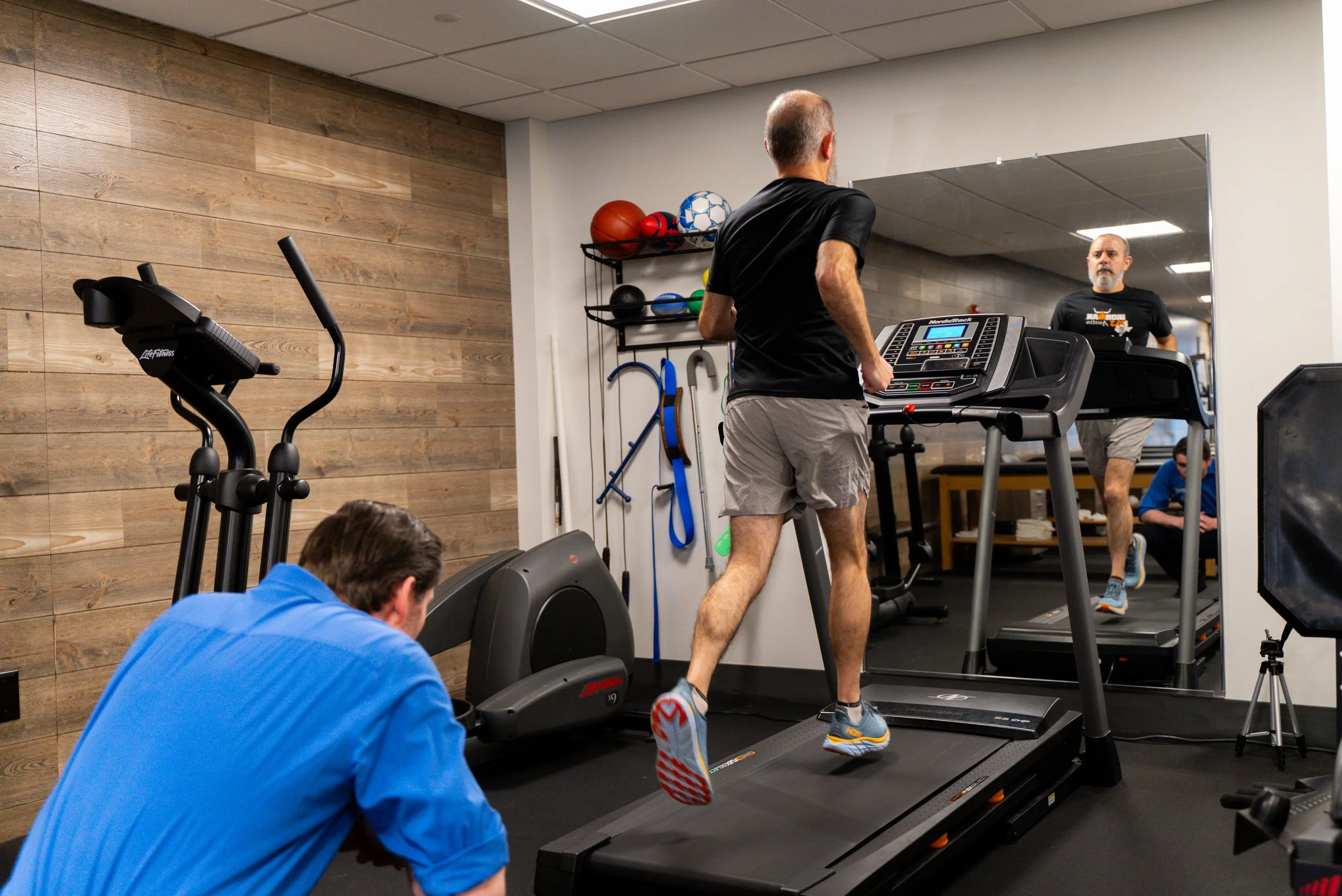 Man jogging on a treadmill in a gym with another person nearby, exercise equipment in the background.