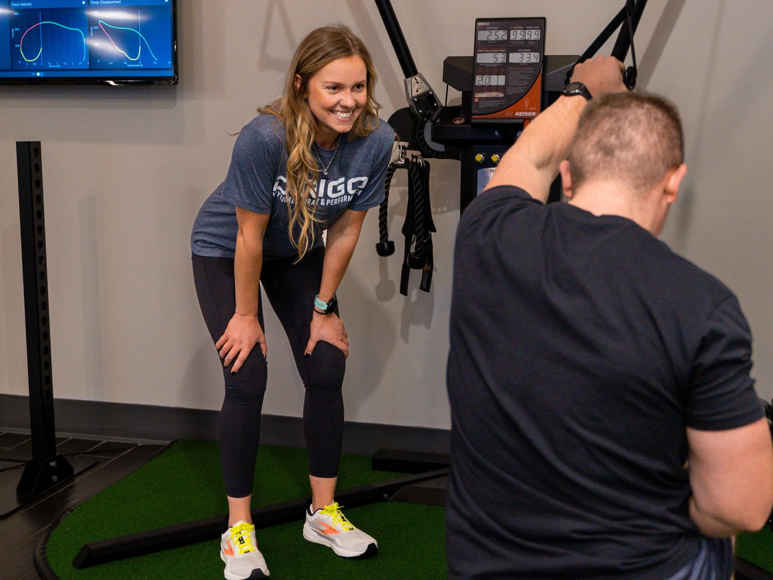 A woman in athletic gear is smiling while standing in a gym, watching a man using exercise equipment. The background includes a screen displaying graphs and workout data.
