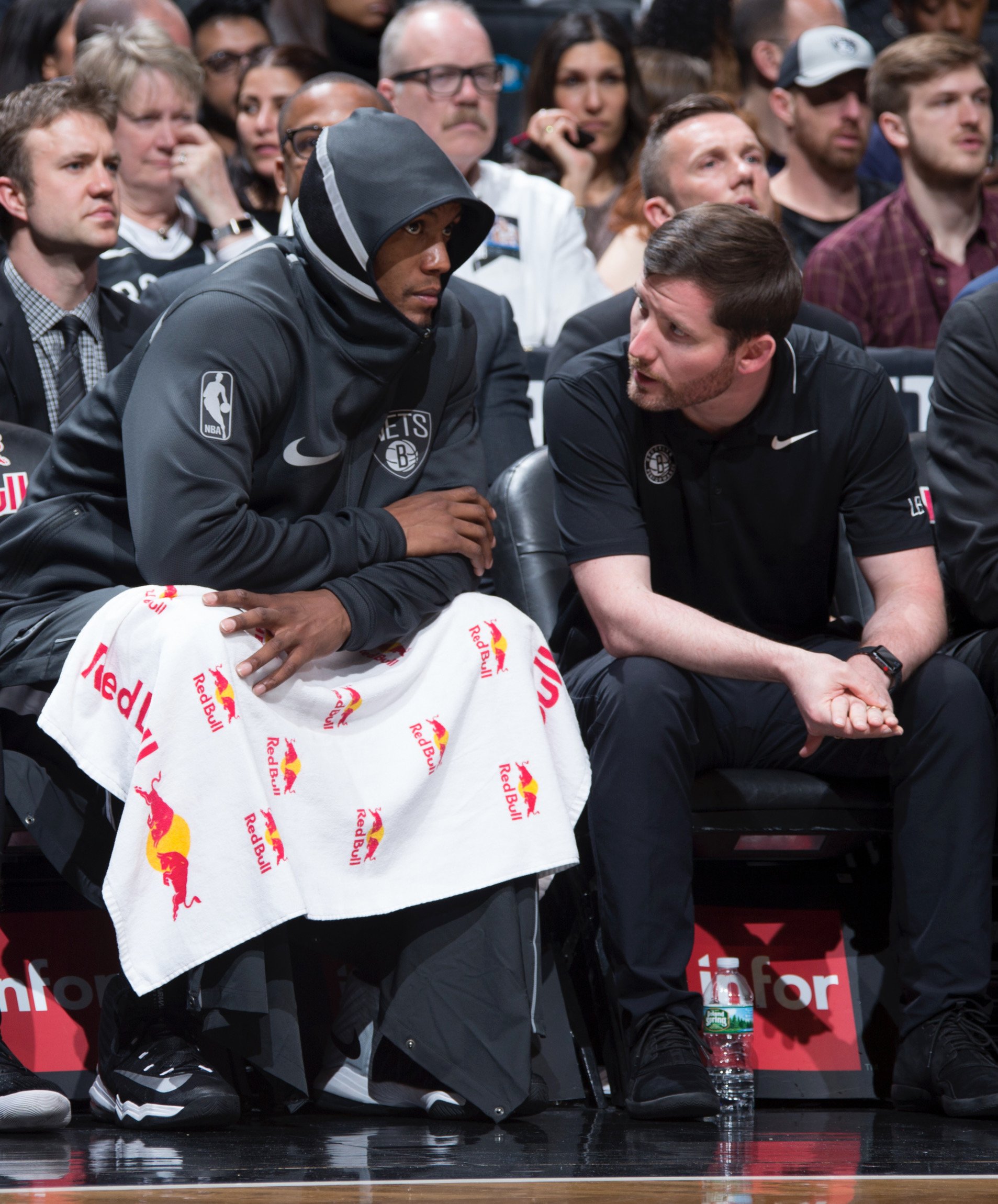 Basketball player in Brooklyn Nets hoodie sitting on bench with coach, draped in Red Bull towel, during a game. Spectators visible in background.
