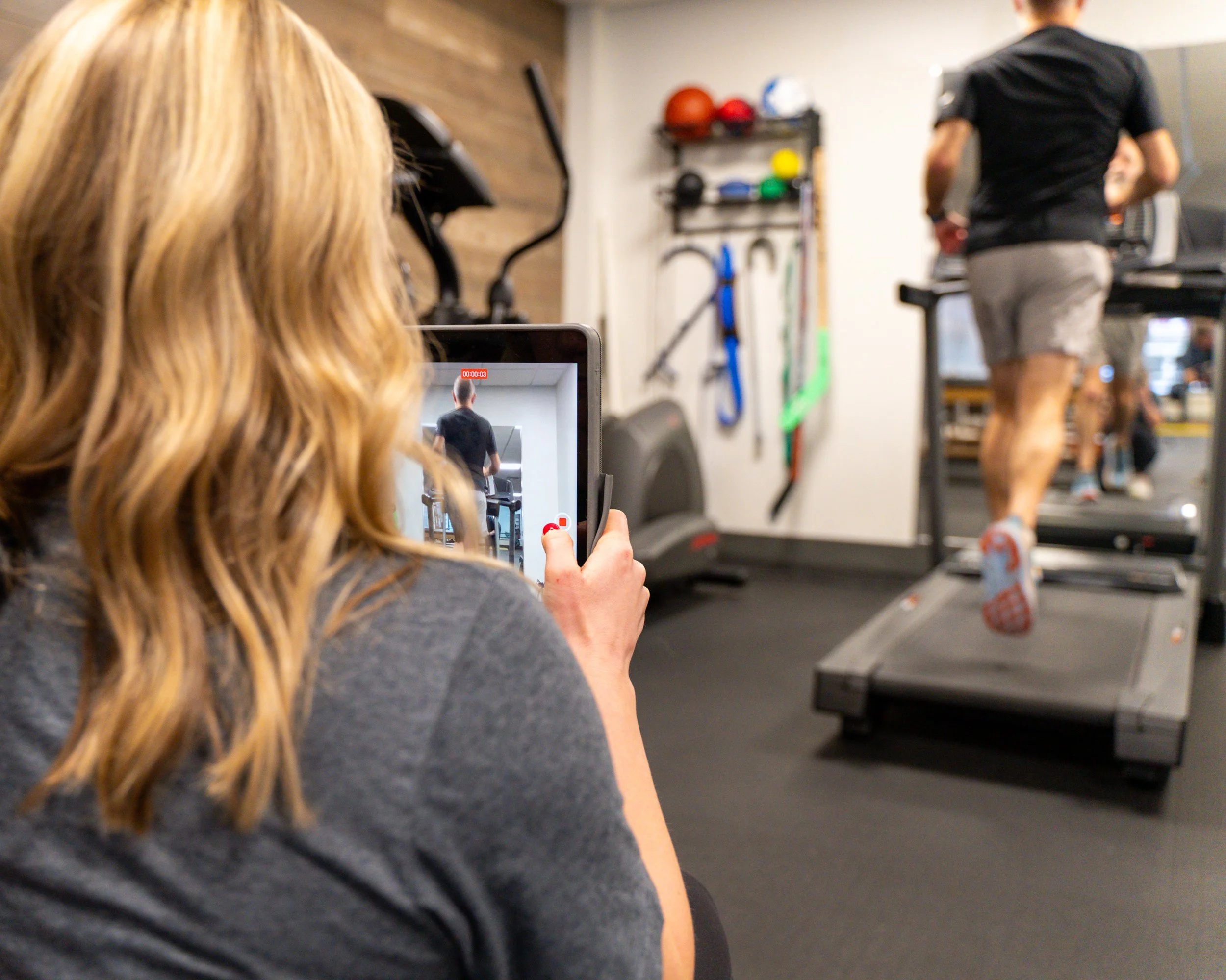 Person recording another person running on a treadmill with a tablet in a gym.