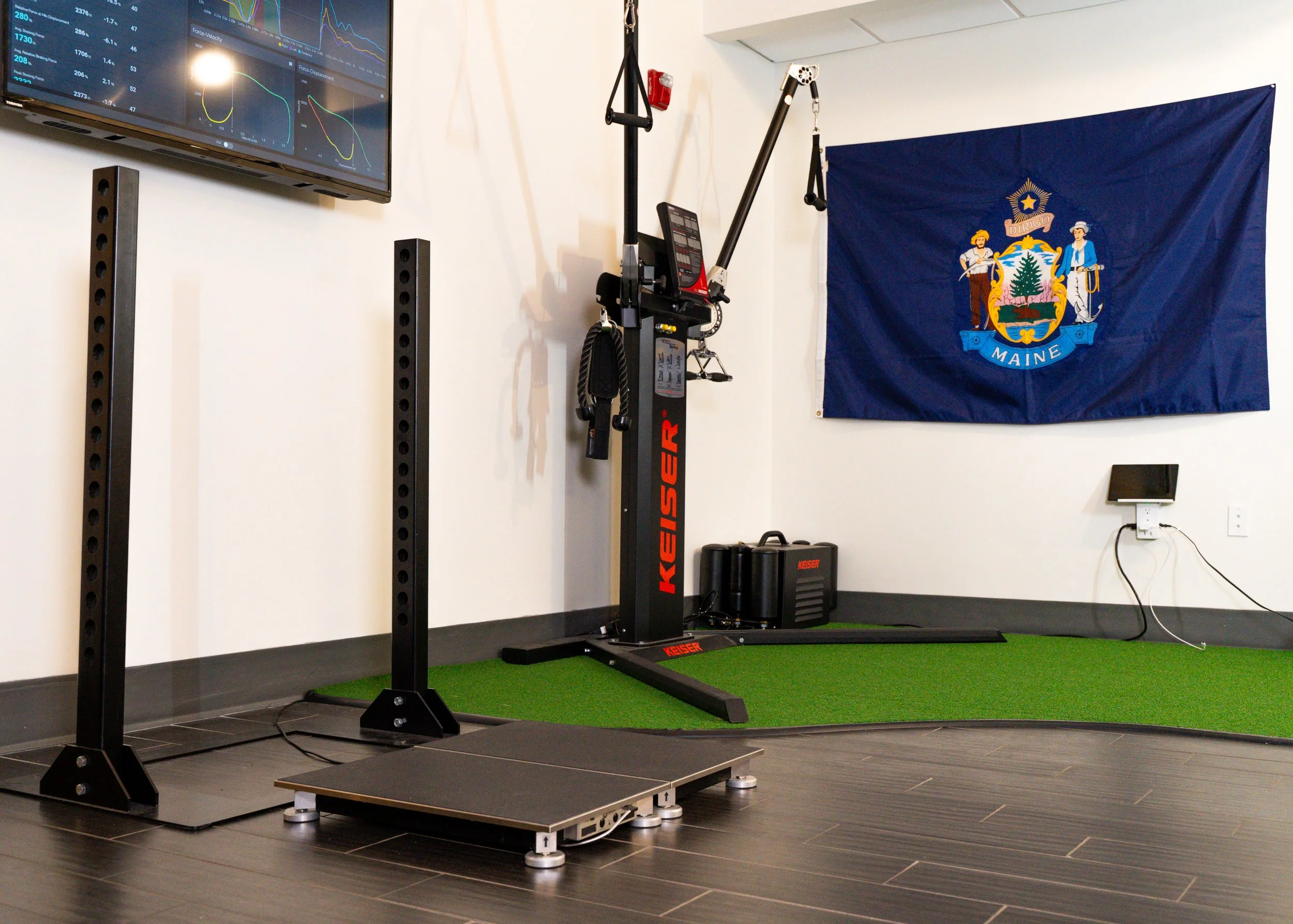 Gym equipment including a Keiser fitness machine and platform, with a Maine state flag in the background.
